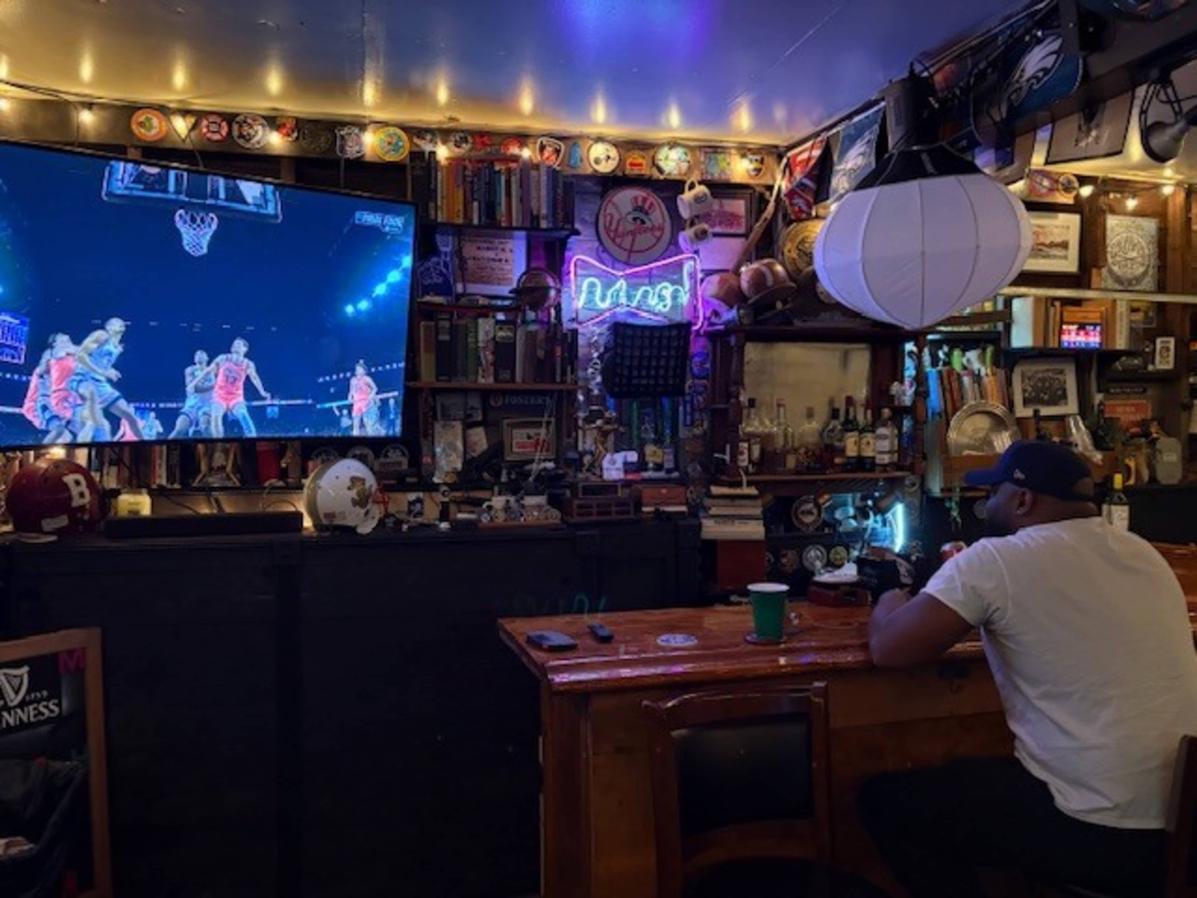 A person sits at a wooden bar watching a basketball game on a large screen in a room filled with memorabilia and dim lighting.