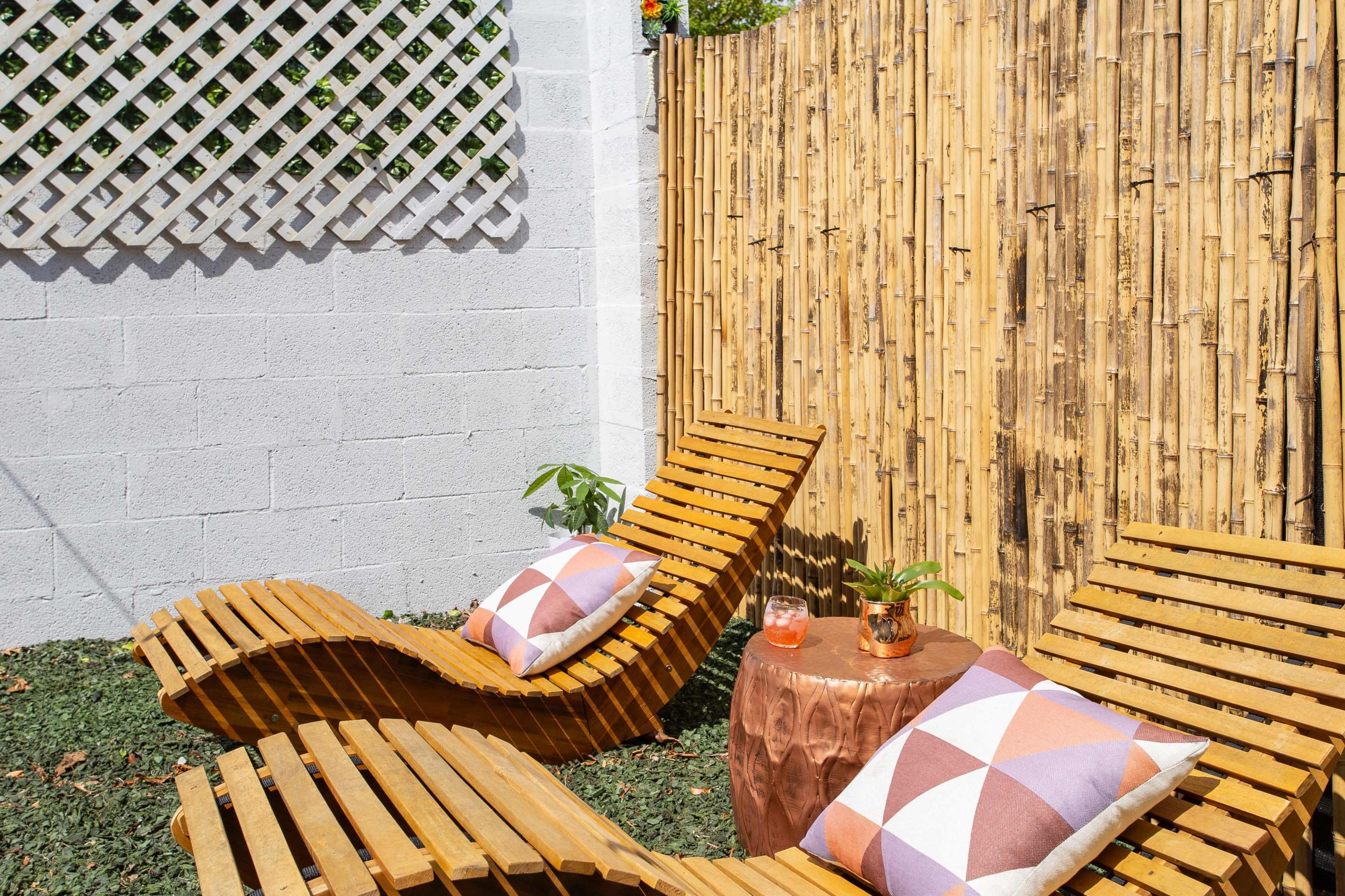 Two wooden lounge chairs with decorative pillows next to a small metal table, surrounded by a bamboo fence and a textured gray wall.
