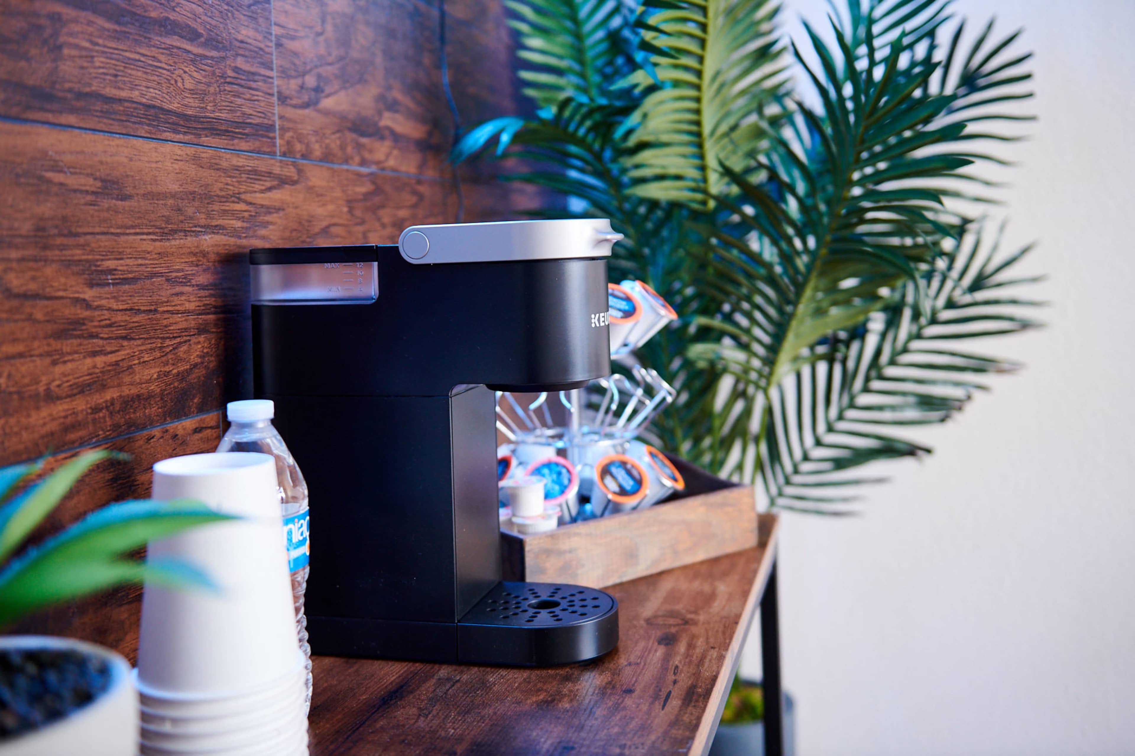 A coffee machine is placed on a wooden table next to a bottle of water and cups, with decorative plants in the background.