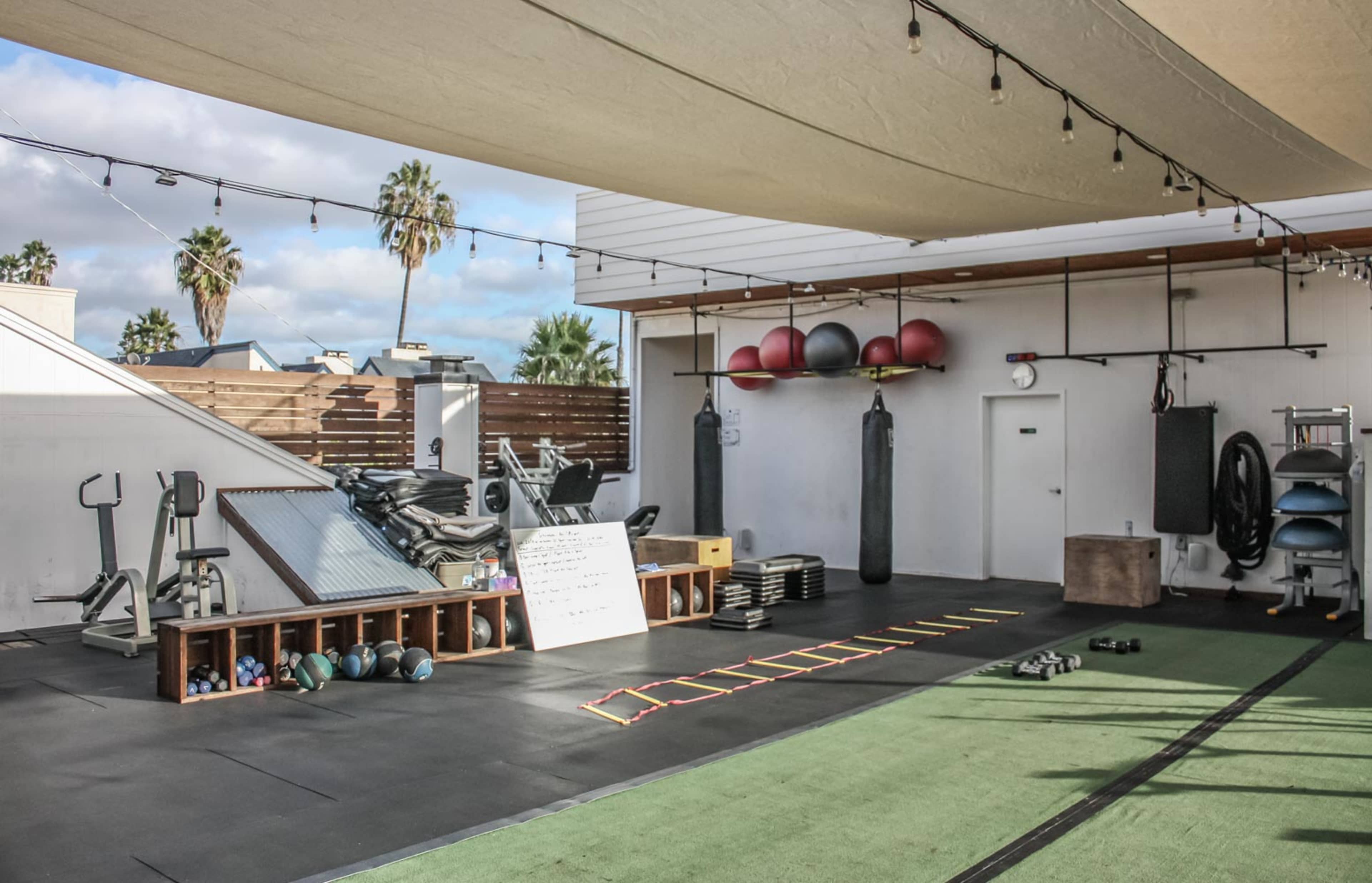 An outdoor gym setup with various exercise equipment, including weights, a mirror, and training tools, beneath a shaded area with palm trees in the background.