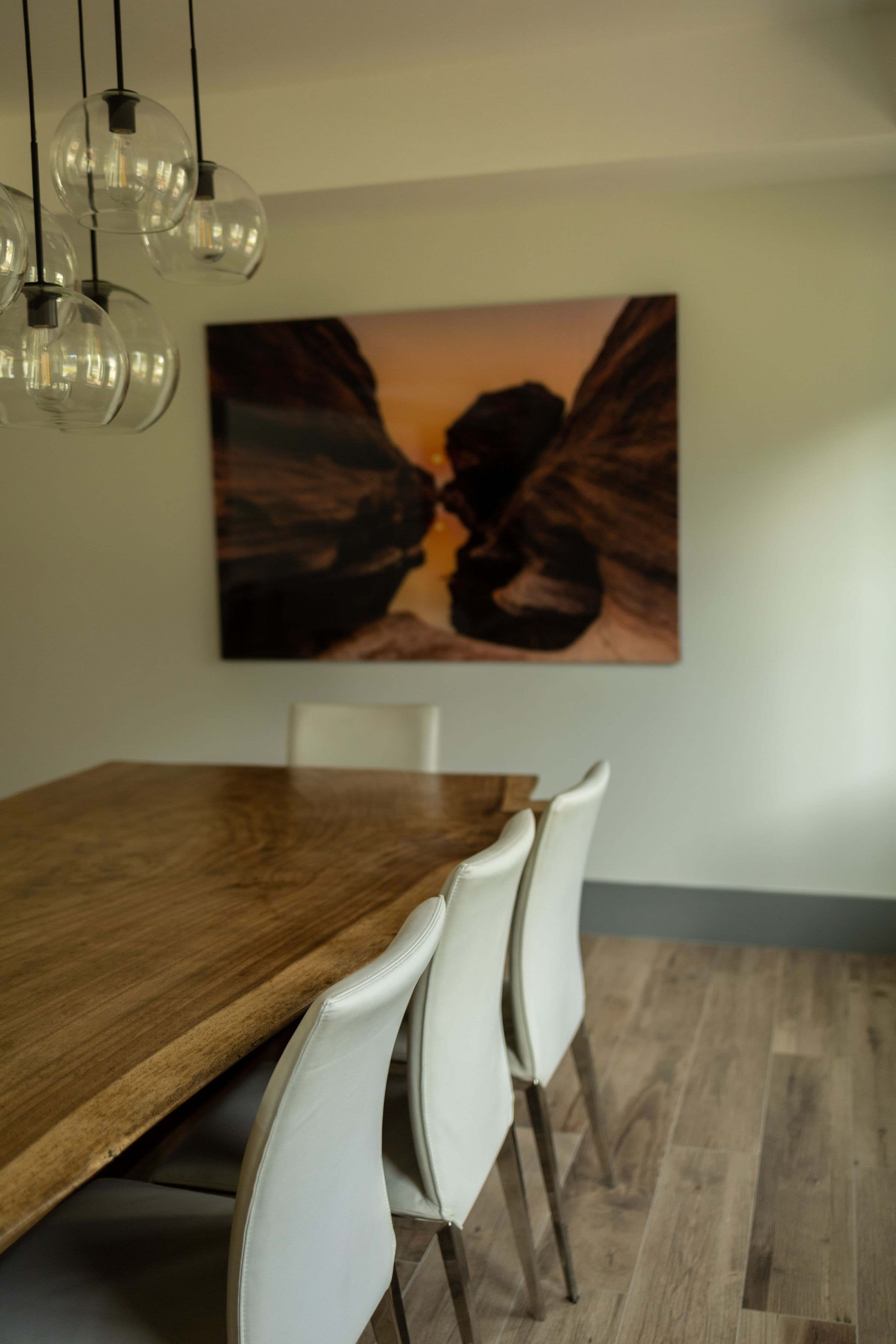 A wooden dining table with four white chairs is positioned beneath a cluster of pendant lights, and a large landscape photograph hangs on the wall behind it.