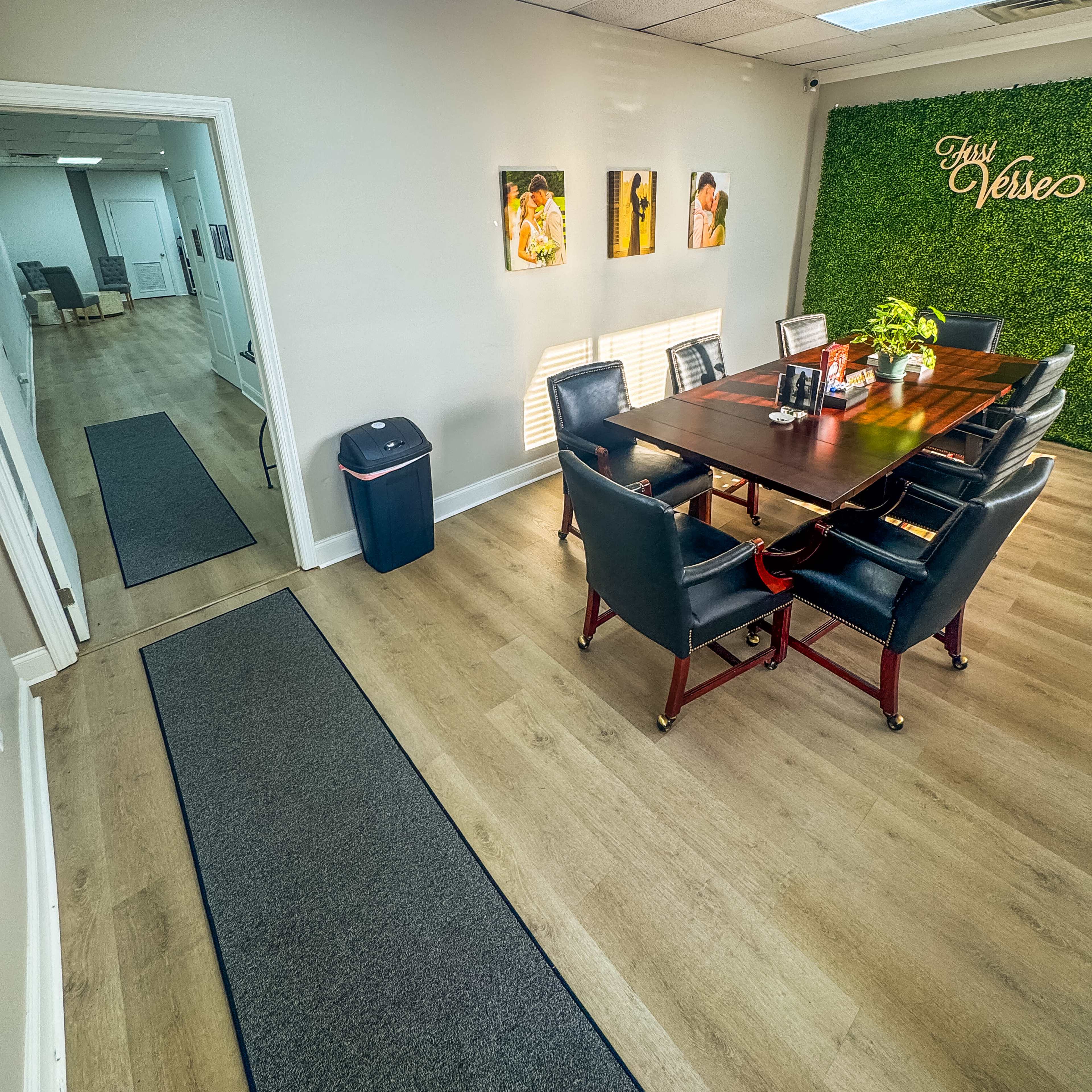 The image shows a conference room with a large wooden table surrounded by black leather chairs, leading to a hallway with a trash can and greenery decor on the wall.