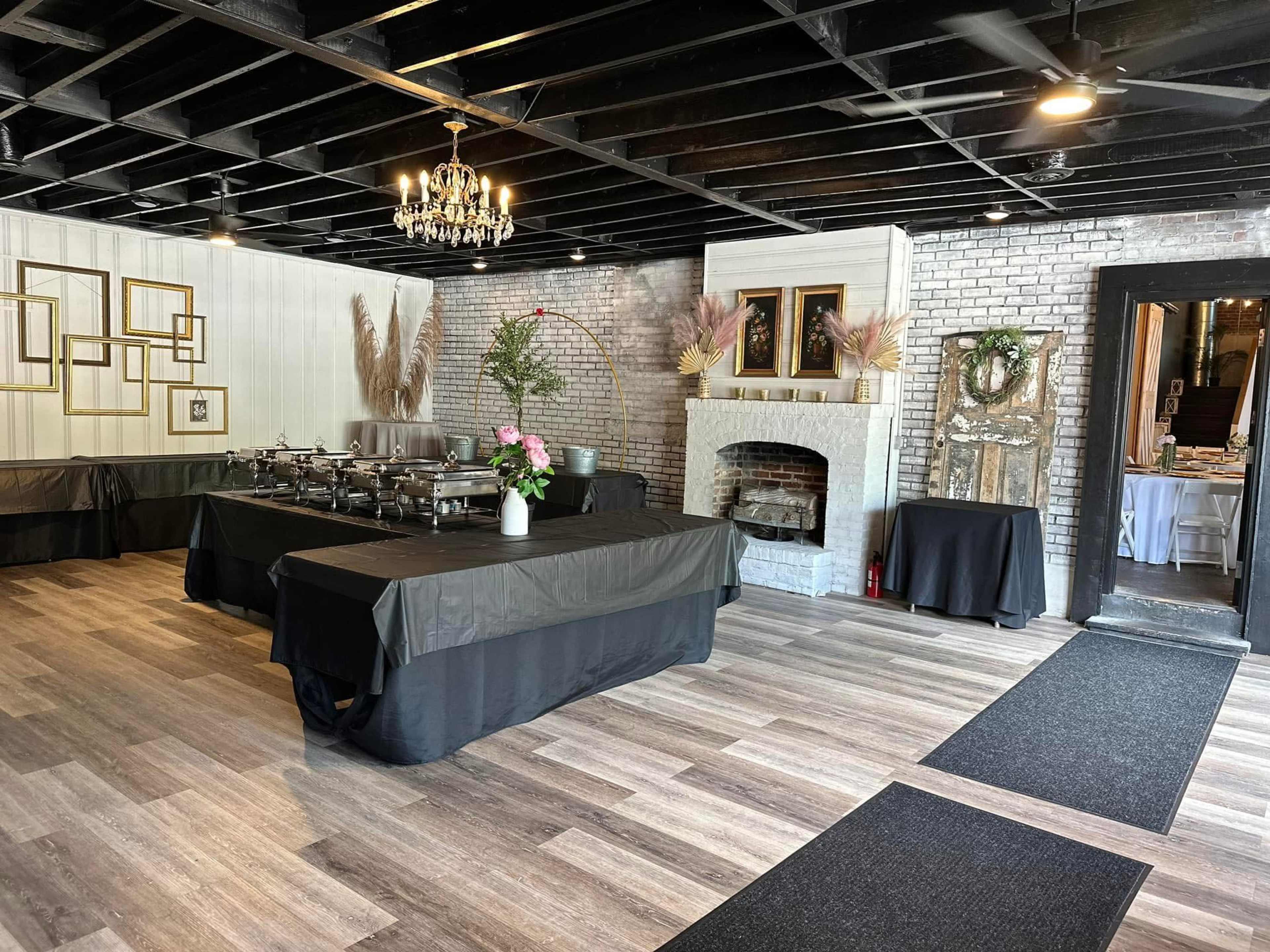 The image shows a spacious event room with a dark ceiling, wooden flooring, and several tables set up for a buffet, along with decorative plants and a fireplace.