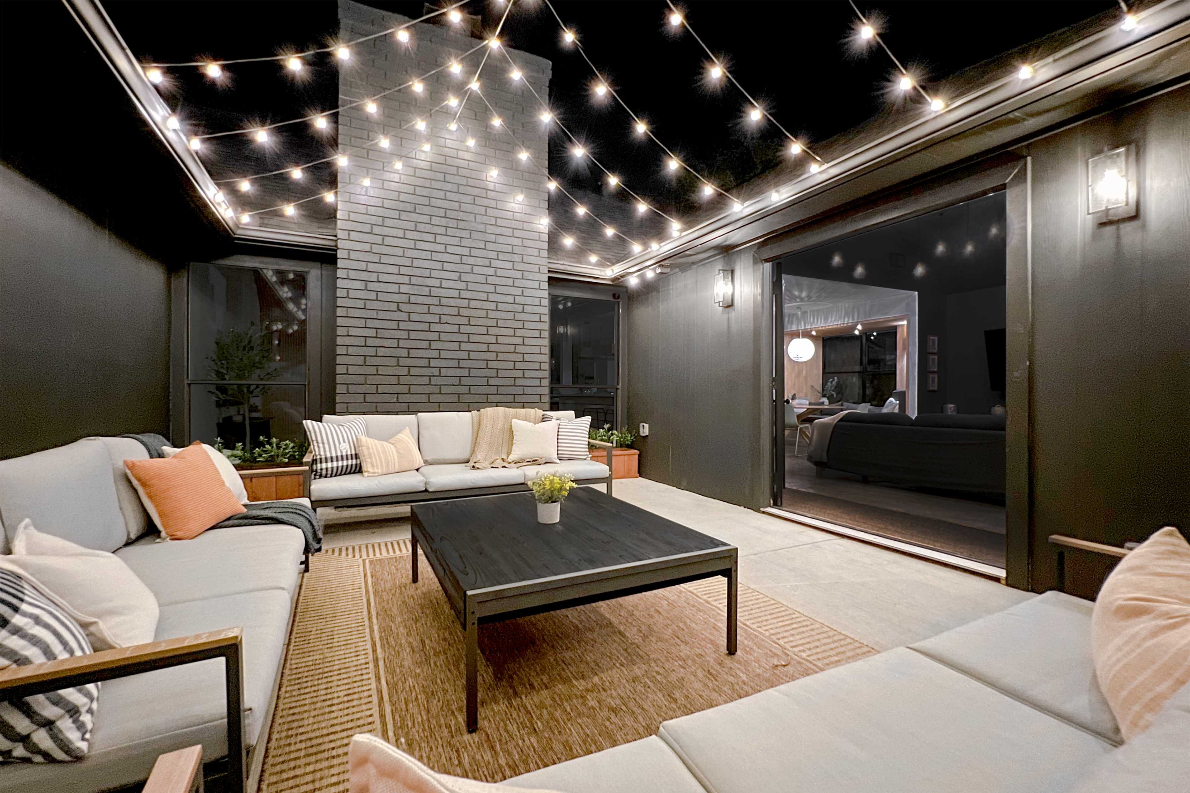 A patio area features cozy seating arranged around a central table, illuminated by string lights above, with a fireplace and an open doorway leading to an indoor space.