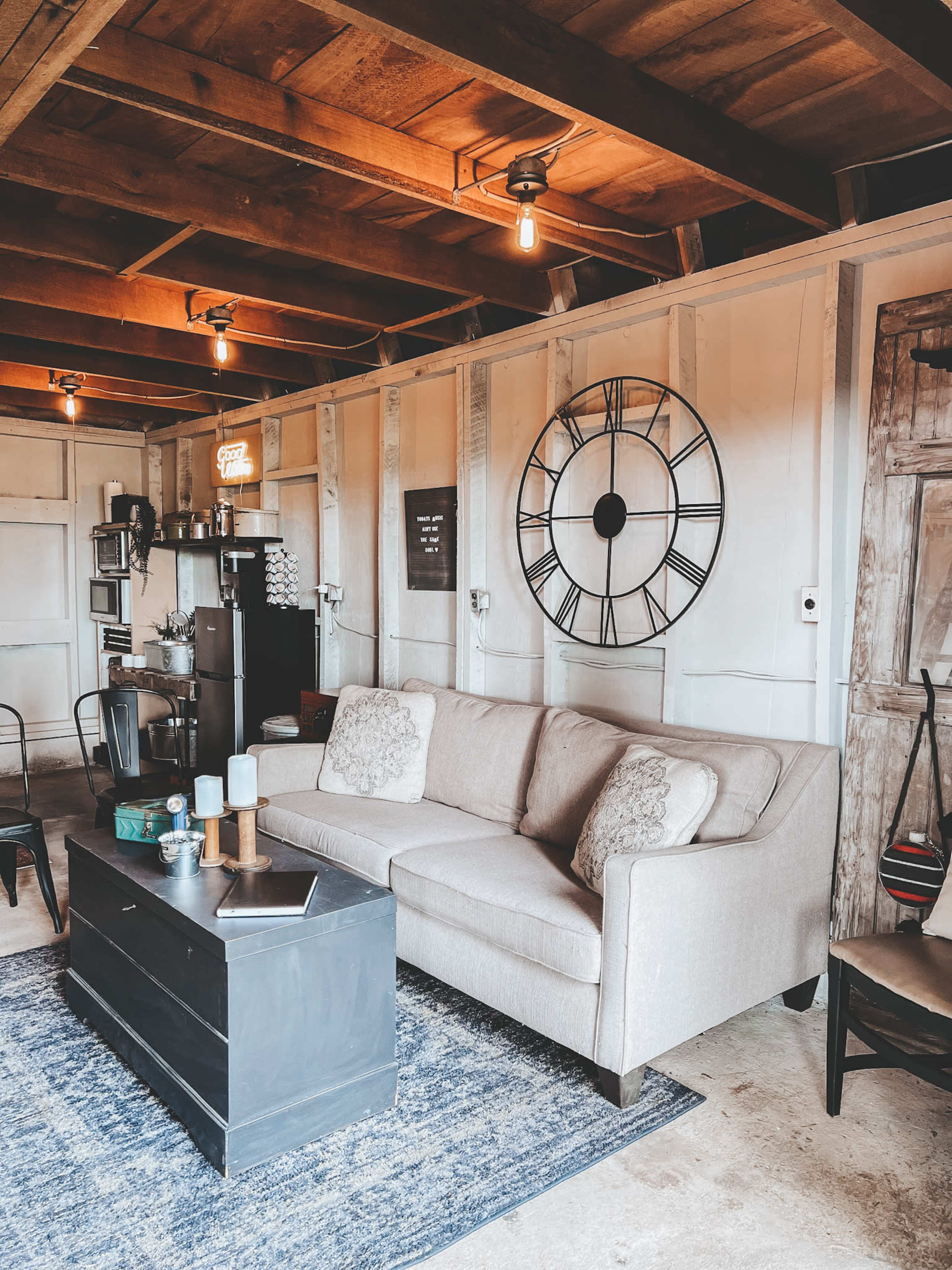 The image depicts a cozy living space featuring a beige sofa, a dark coffee table, and a large wall clock against a paneled wall.