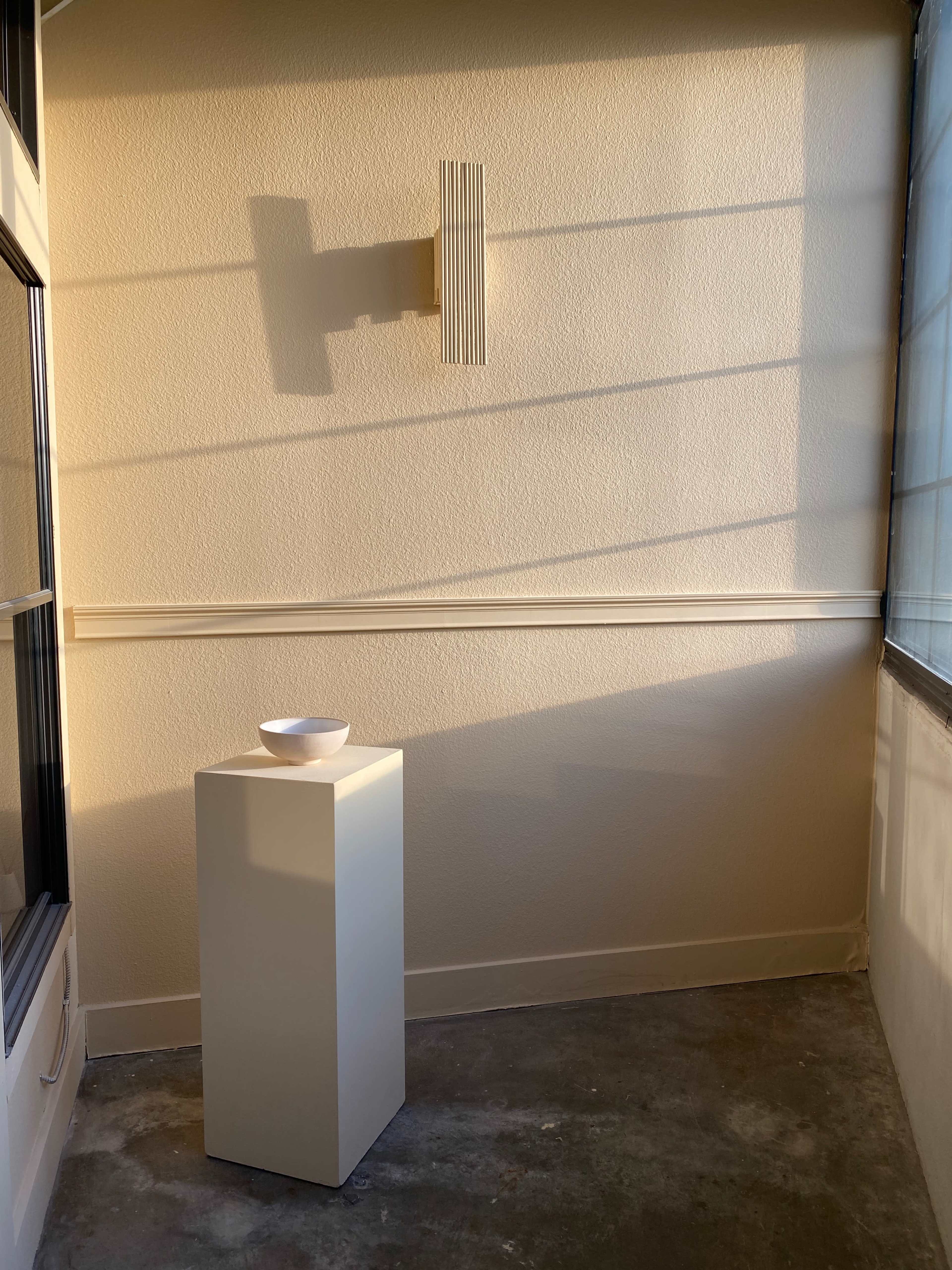 A white pedestal supports a bowl in a softly lit corner of a room with beige walls and a decorative wall fixture.