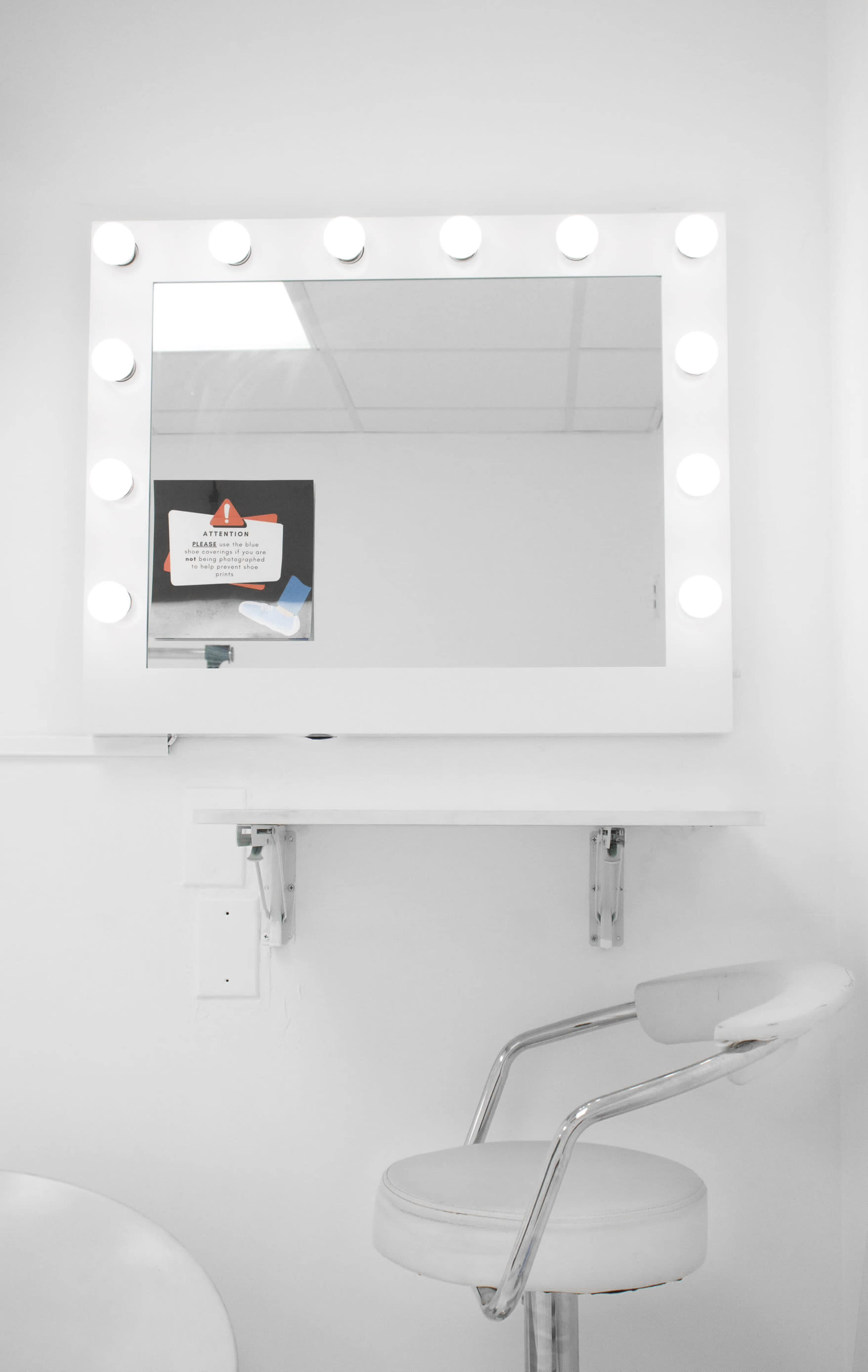 A well-lit vanity mirror with a white frame and circular lights is mounted above a small shelf and a chrome stool.