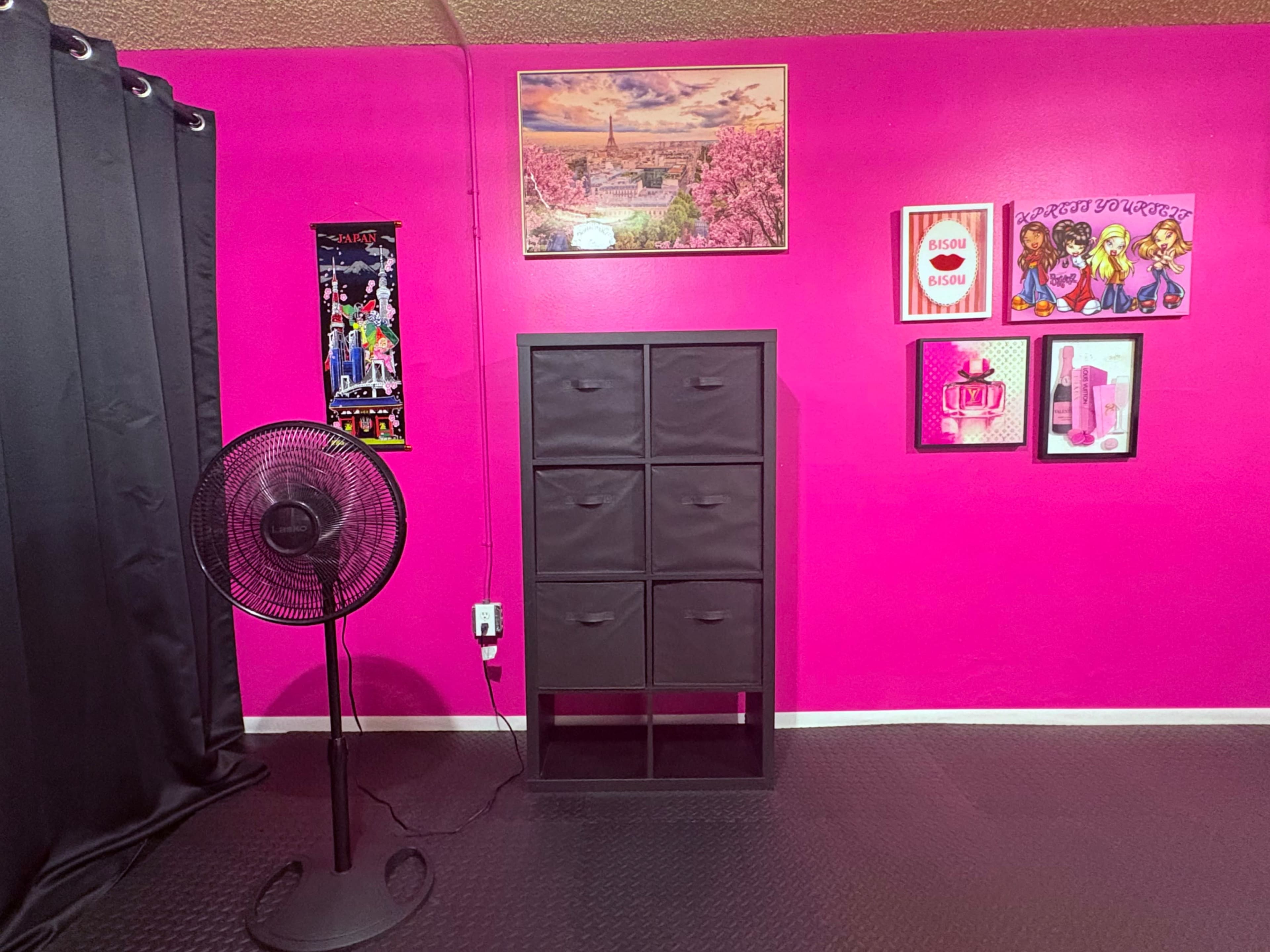 The image features a bright pink room with a black fan and a black storage cabinet, surrounded by various framed artworks on the walls.
