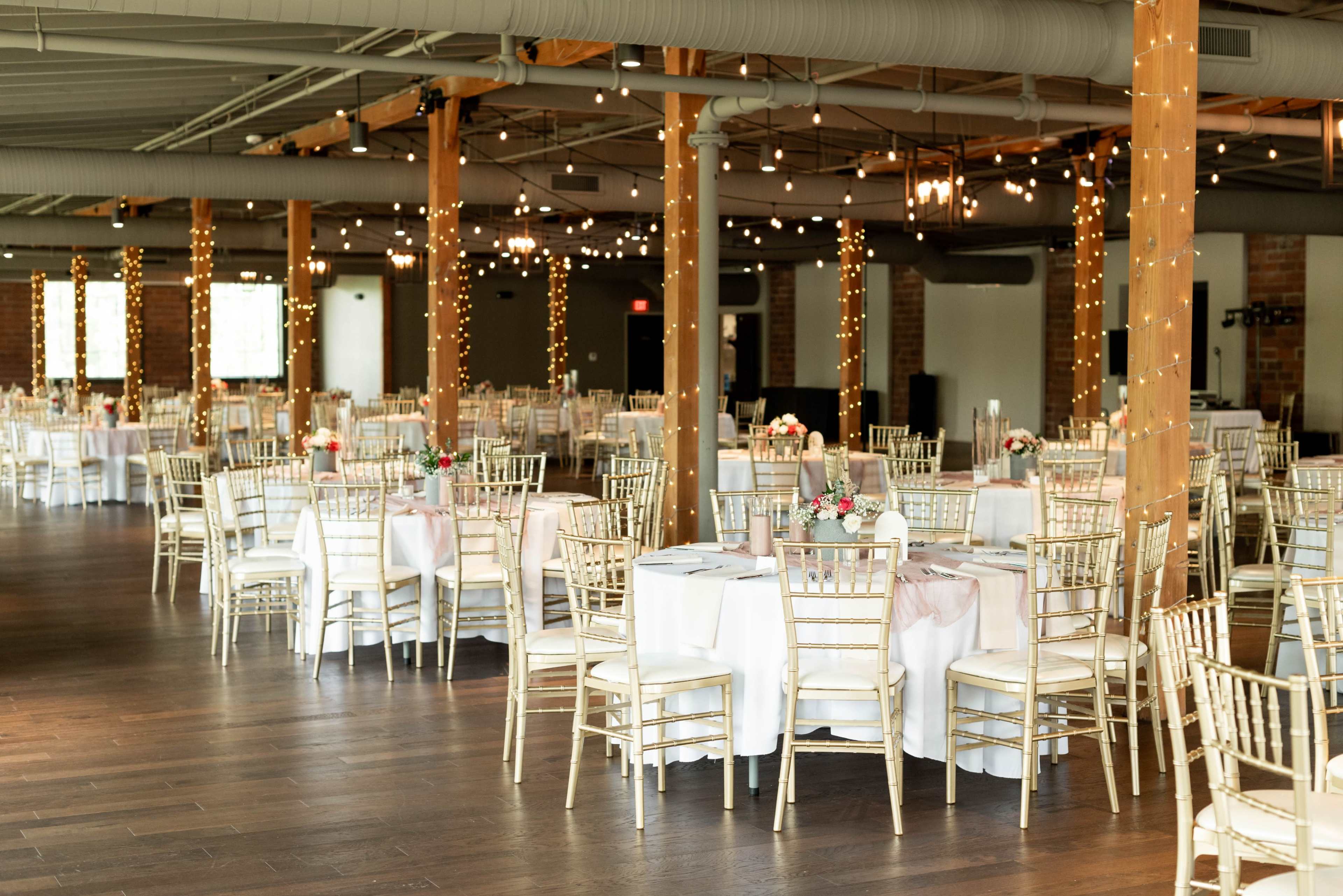 A spacious event venue features elegantly set tables with white tablecloths and gold chiavari chairs, illuminated by string lights and natural light from large windows.