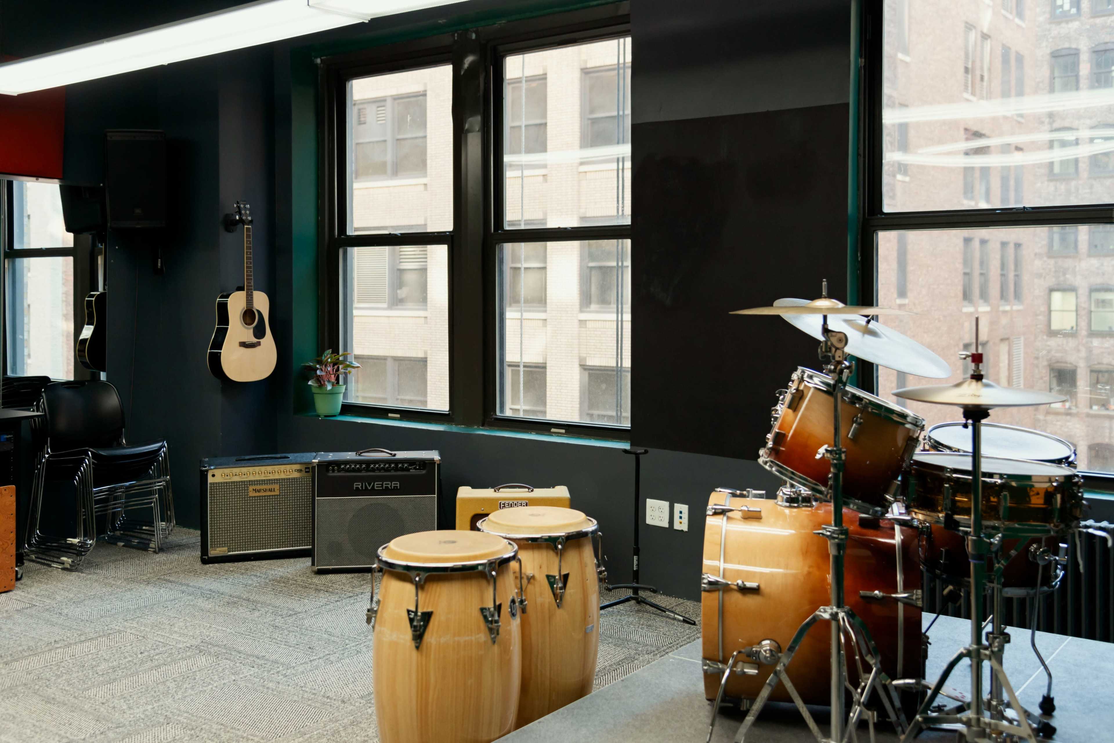 The image shows a music practice room featuring a drum set, congas, amplifiers, a guitar on the wall, and large windows overlooking a cityscape.