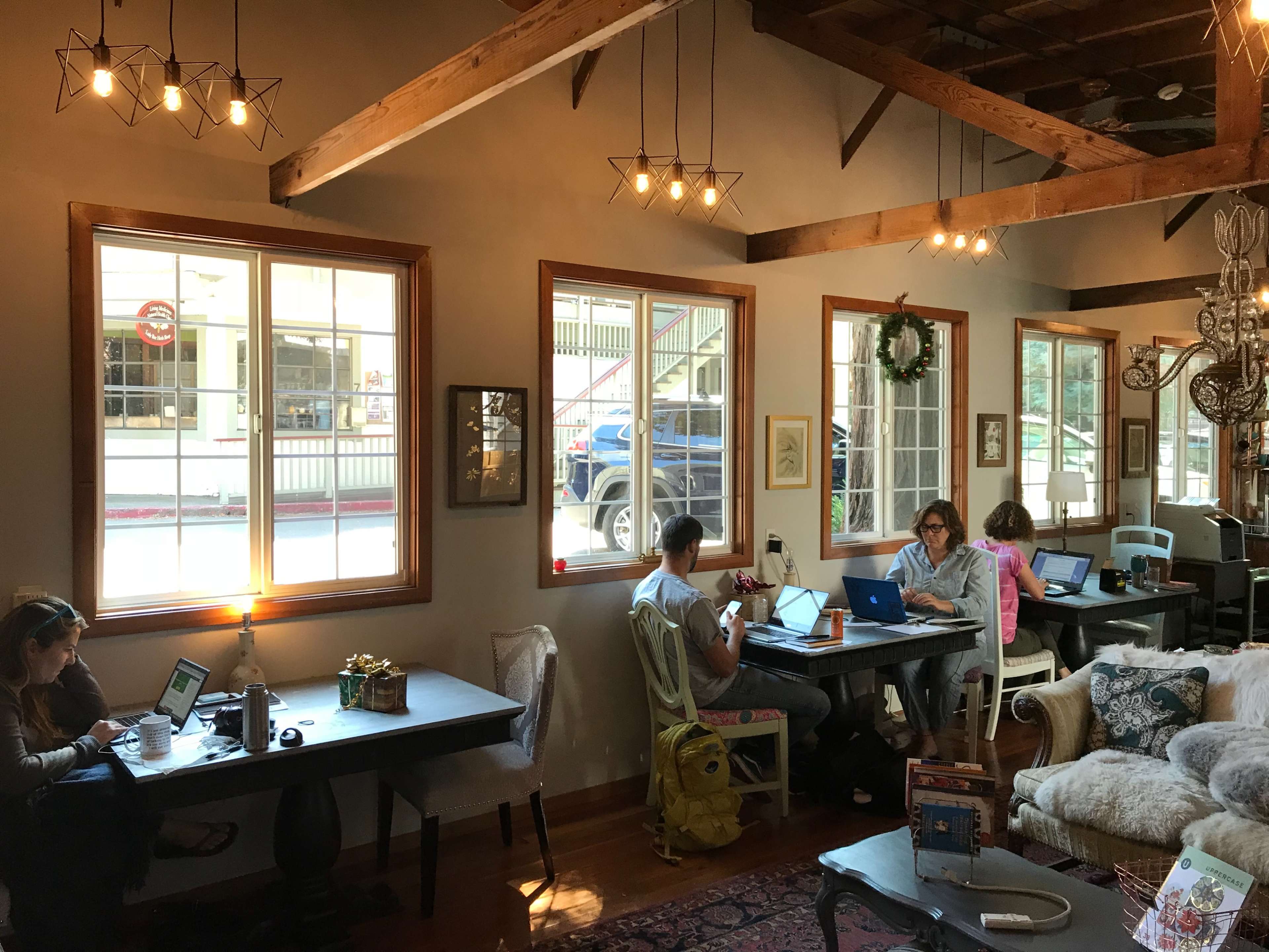 A cozy cafe interior with several individuals working on laptops at wooden tables near large windows.