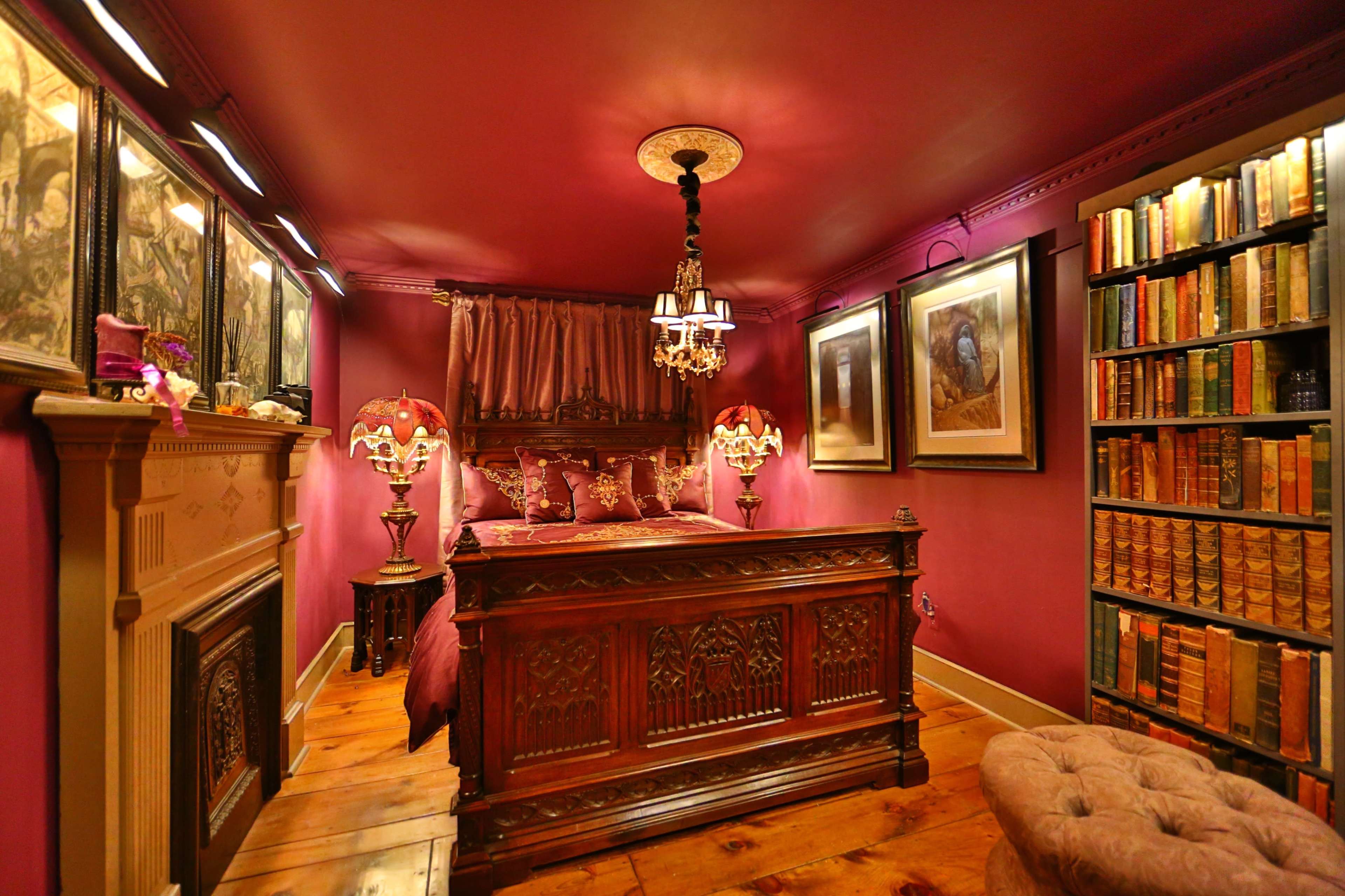The image shows a small, elegantly furnished bedroom with a wooden bed, antique lamps, a bookcase filled with books, and decorative artwork on the walls.