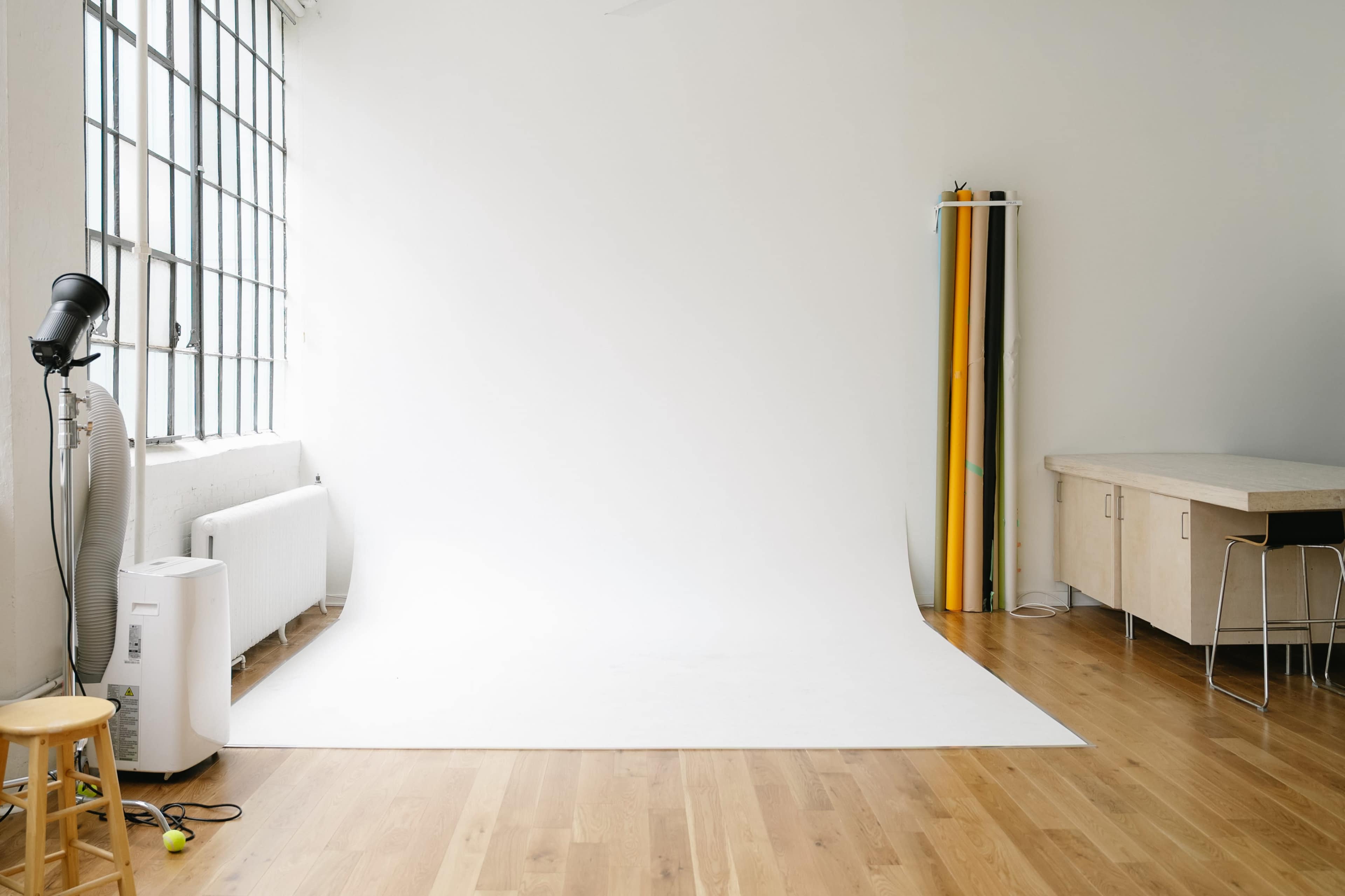 The image shows a bright, empty photo studio with a white backdrop and a few rolls of colored paper against the wall.