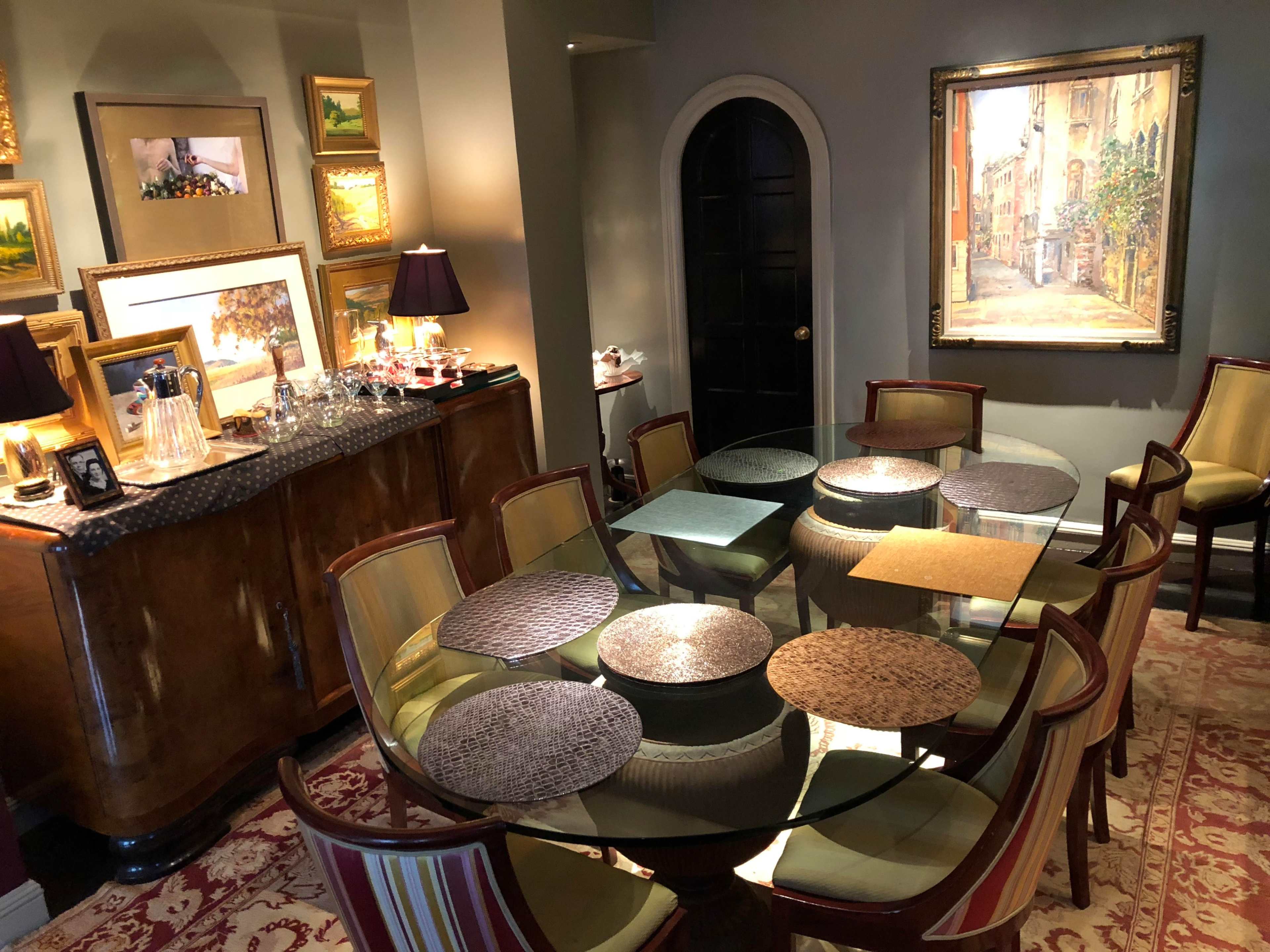 A dining area features a circular glass table surrounded by twelve chairs, with an ornate cabinet holding glassware and artwork adorning the walls.