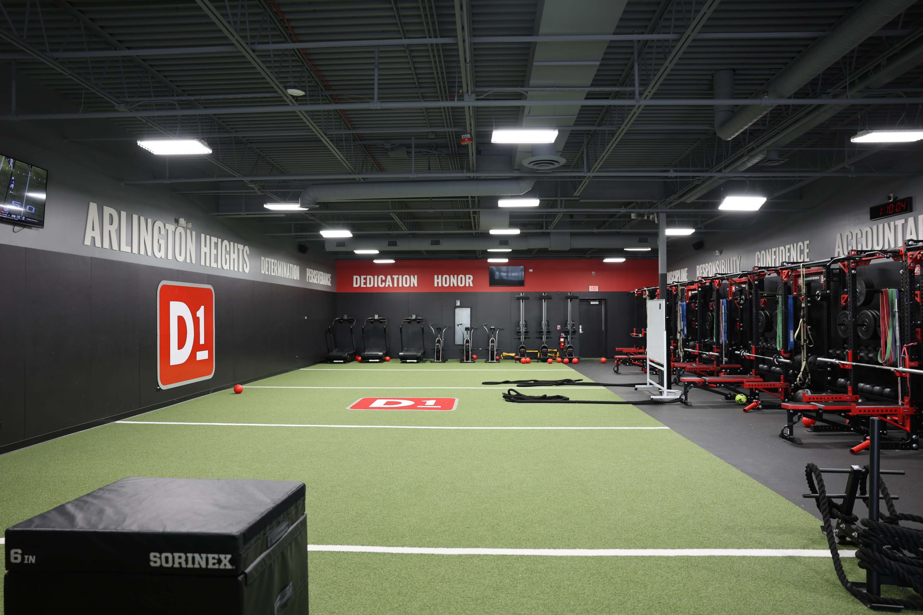 The image shows a training facility with artificial turf, equipped with various fitness equipment and workout stations along the walls, and "D1" branding prominently displayed.