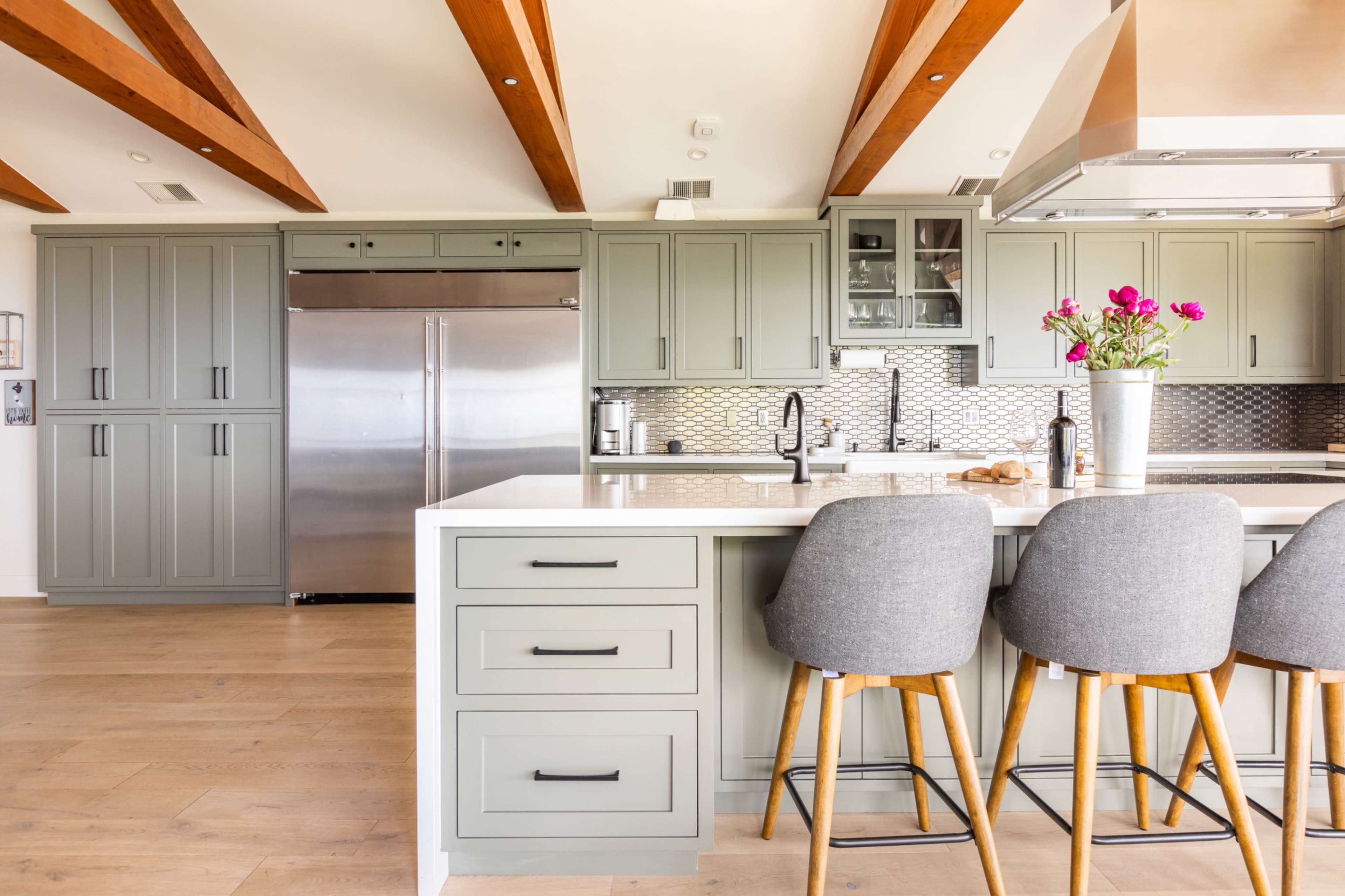 The image shows a modern kitchen with green cabinetry, stainless steel appliances, and a white countertop with three gray bar stools.