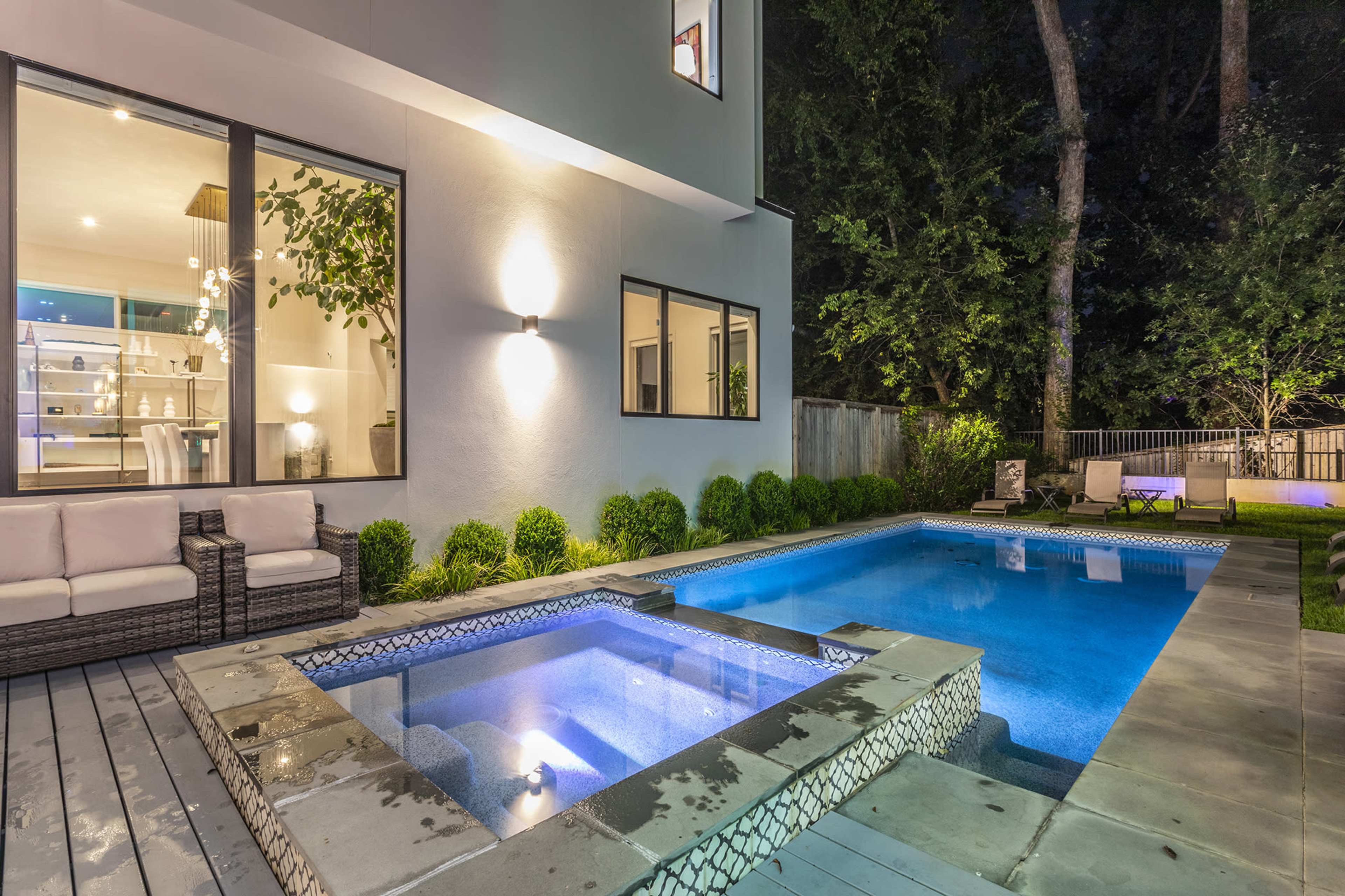 A modern backyard features a swimming pool and a separate spa, illuminated under nighttime lighting, with seating and landscaped greenery nearby.
