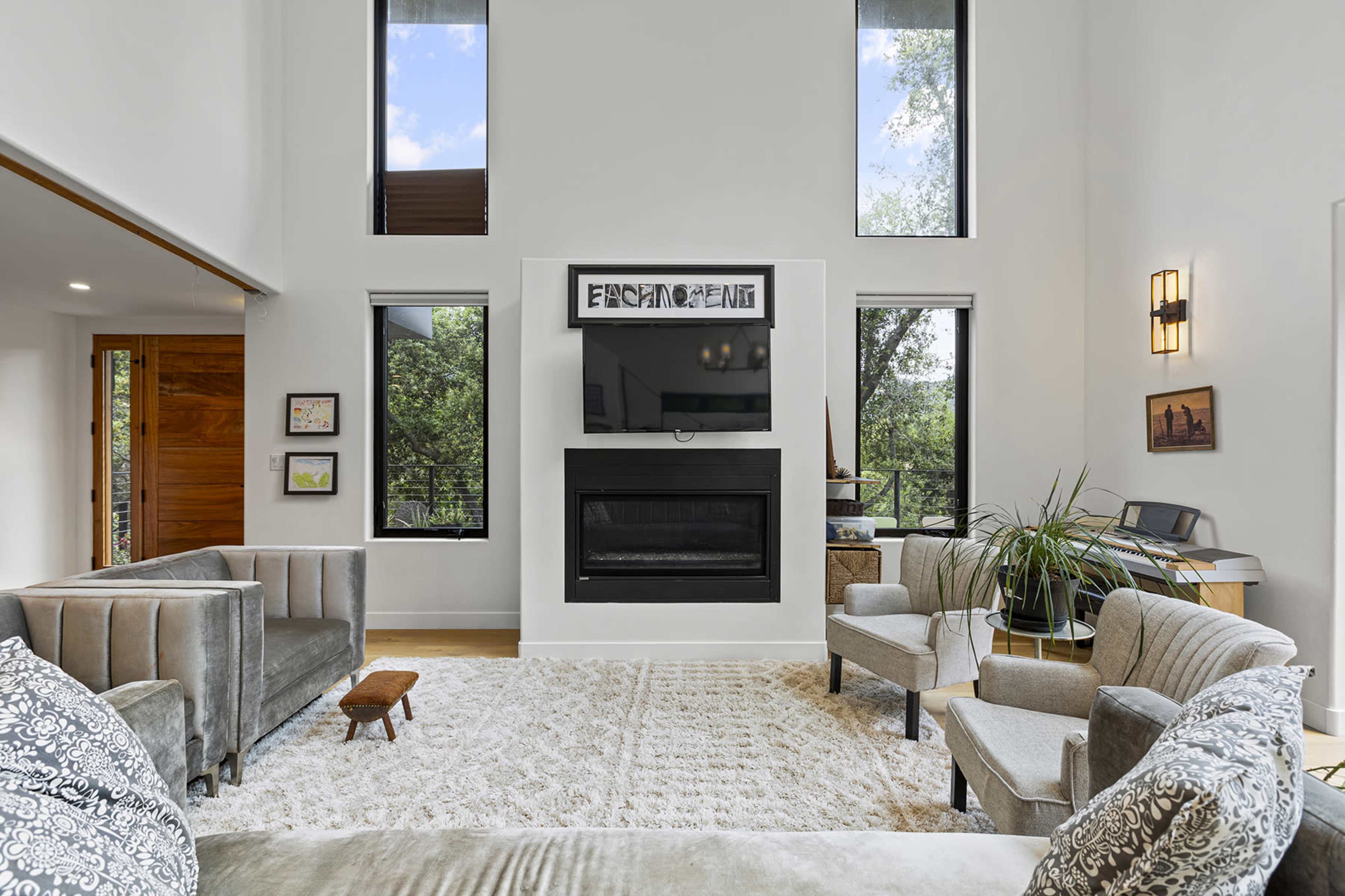 The living room features a fireplace centered on a white wall, flanked by large windows that allow natural light to illuminate the space, furnished with gray sofas and armchairs on a light rug.