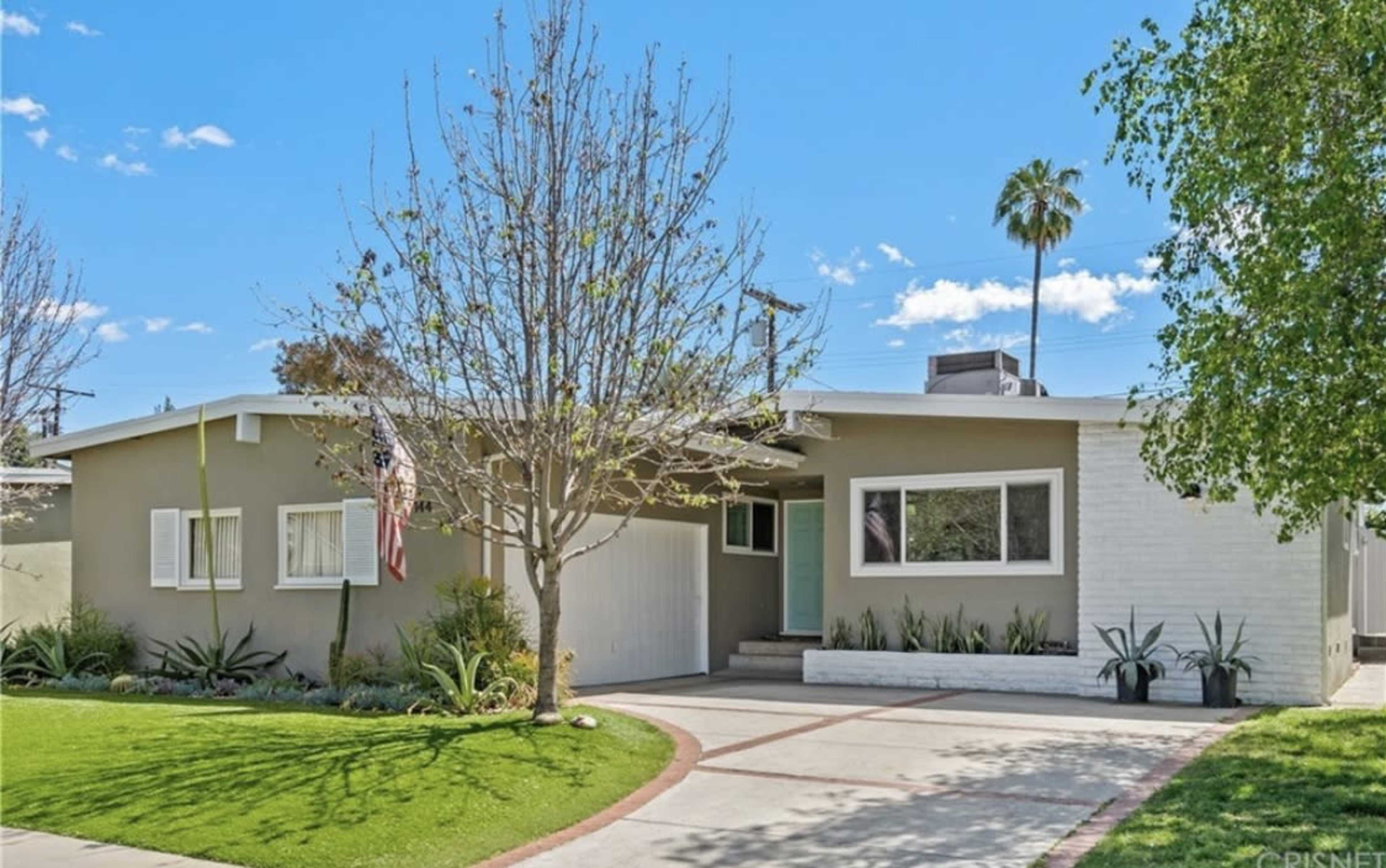 The image shows a modern single-story house with a landscaped front yard, featuring a concrete driveway and a flagpole.