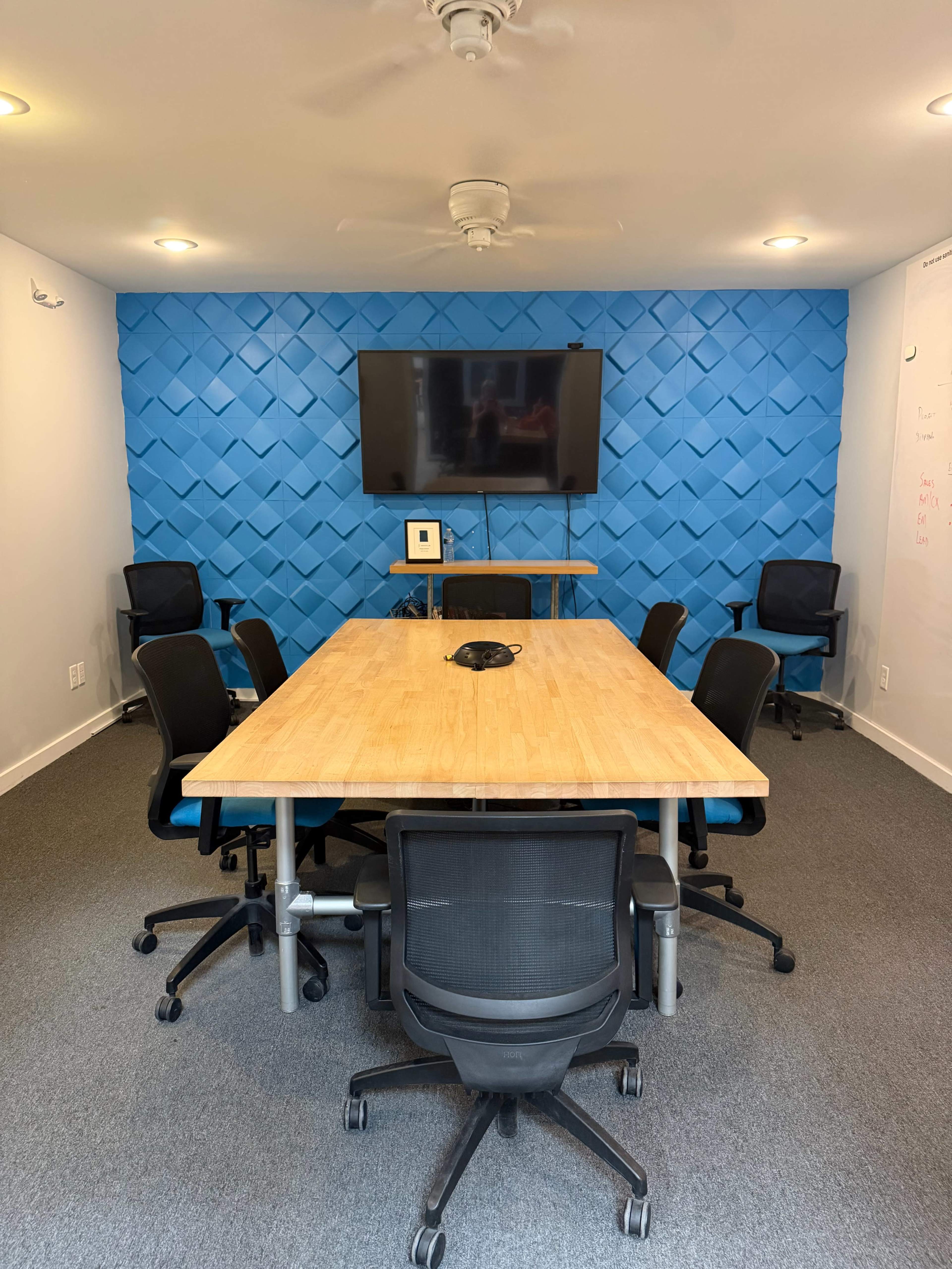 A conference room features a large wooden table surrounded by black chairs, with a blue accent wall and a mounted television at one end.