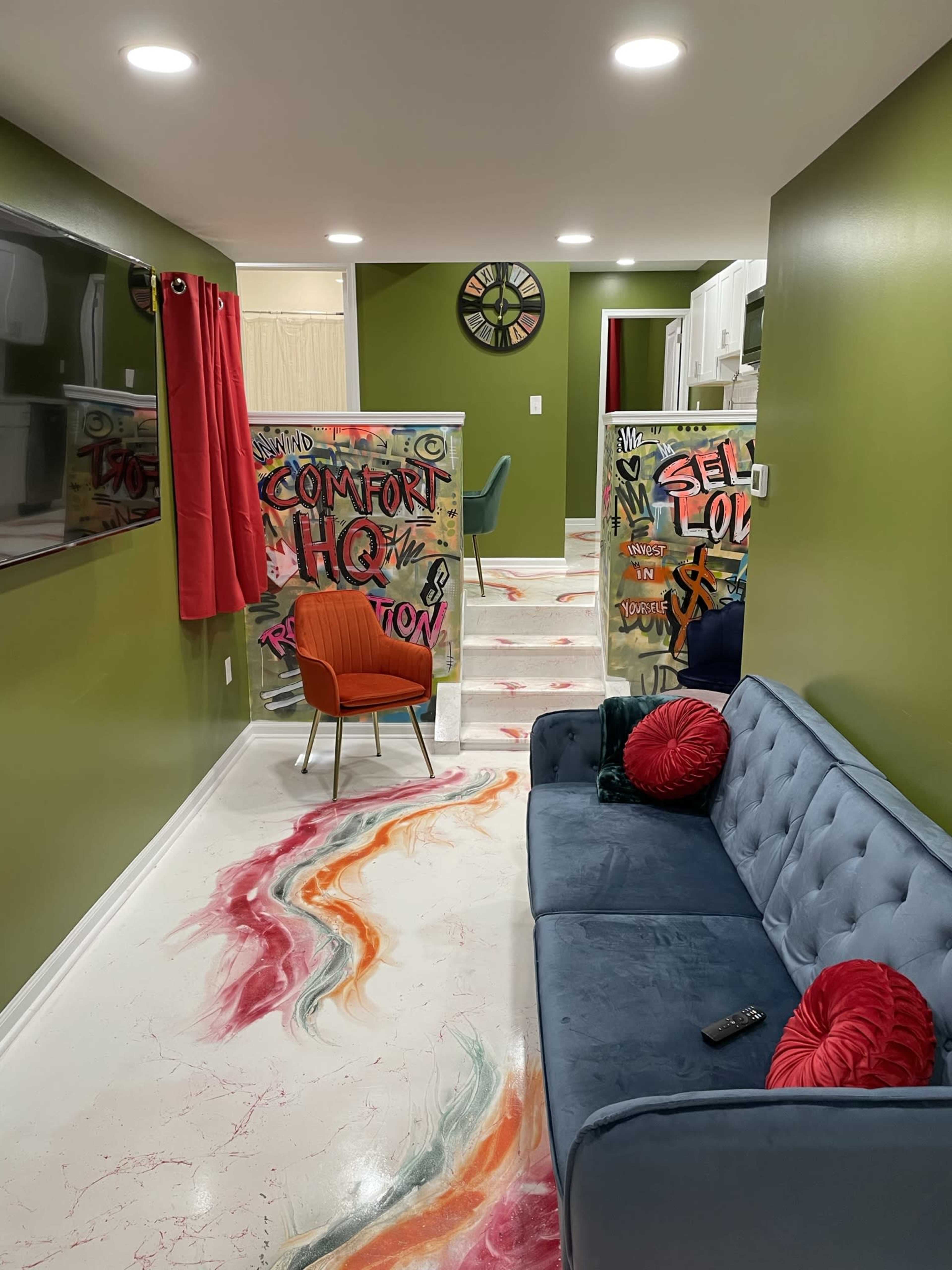 A modern living space with green walls, a blue sofa, colorful graffiti on partitions, and a patterned floor leading to a kitchen area.