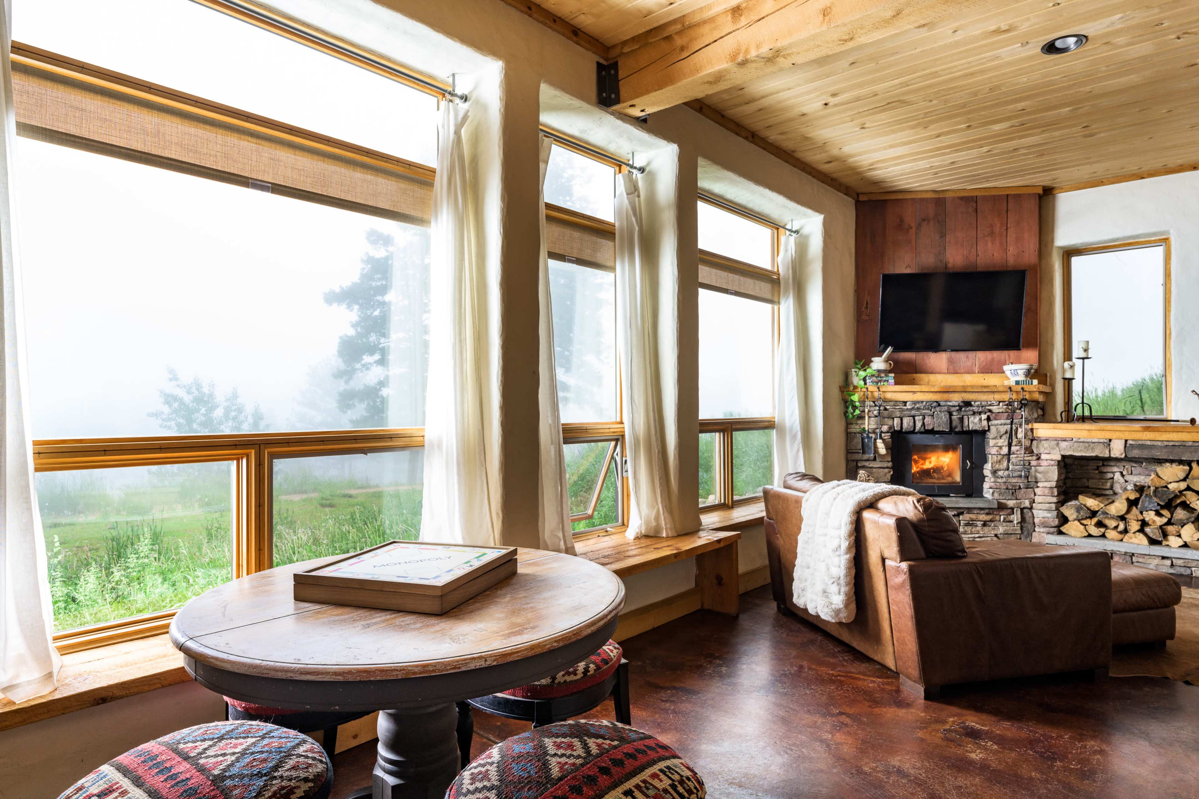 The room features large windows with a view of a foggy landscape, a stone fireplace, and a round table with patterned chairs.