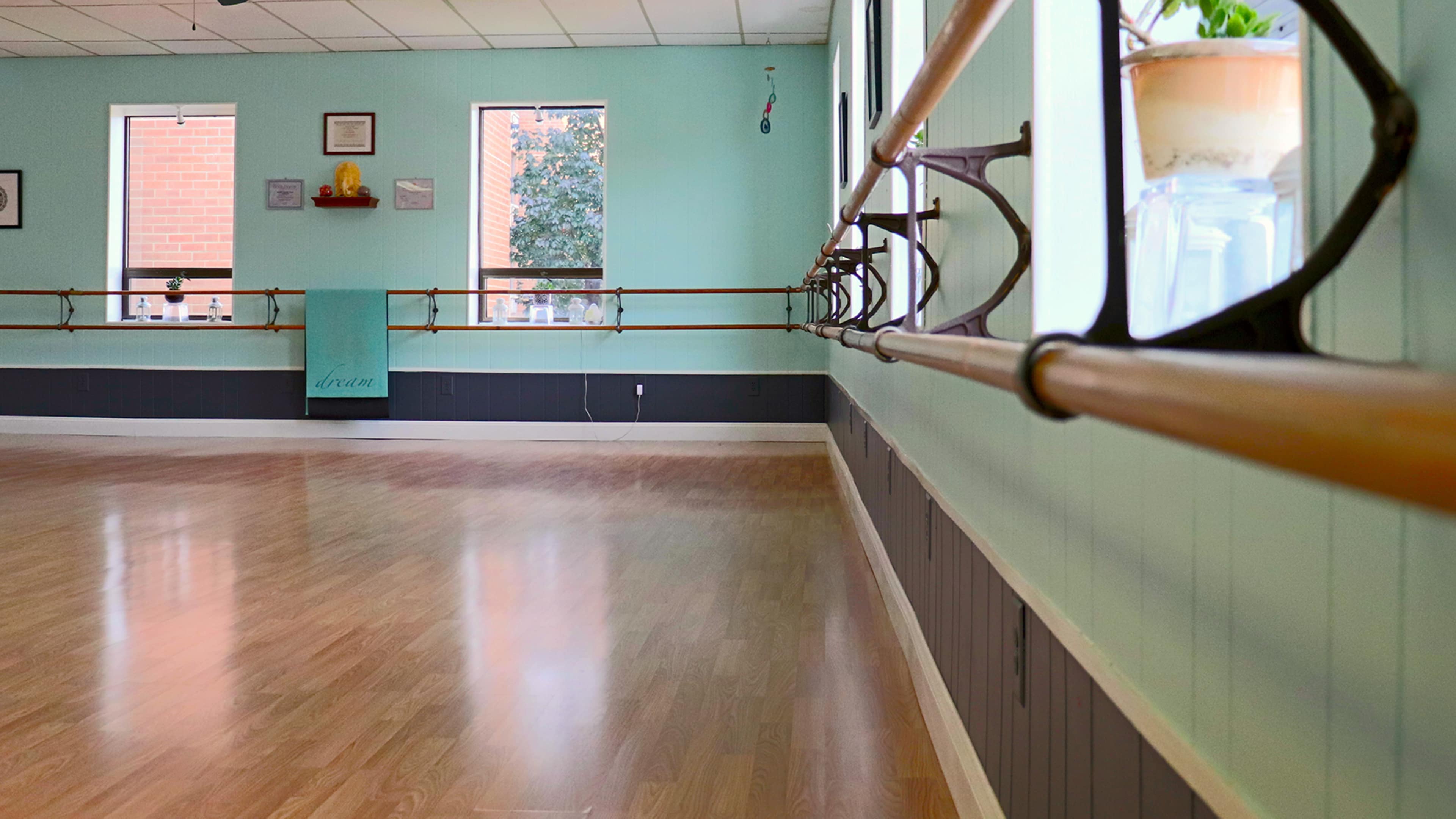 The image shows a dance studio with wooden ballet bars, light-colored walls, and large windows allowing natural light to enter.
