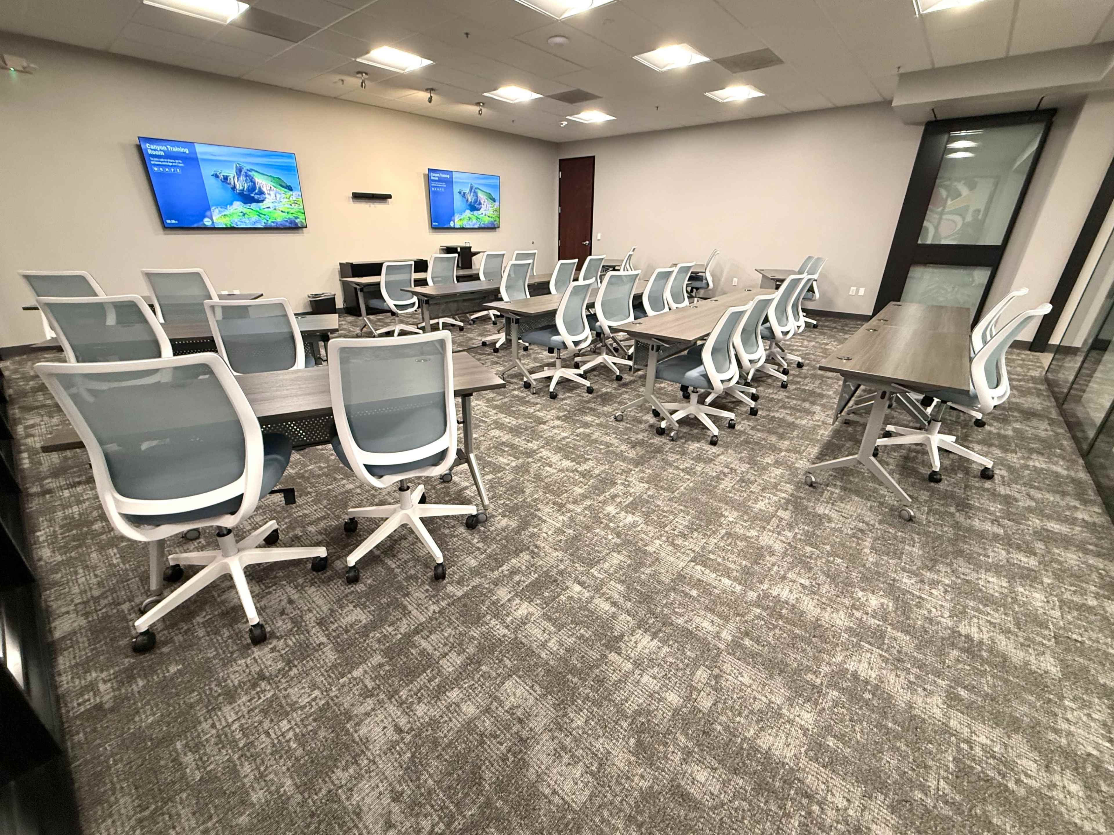 The image shows a modern conference room with multiple tables and chairs arranged for a meeting, featuring two screens mounted on the walls.