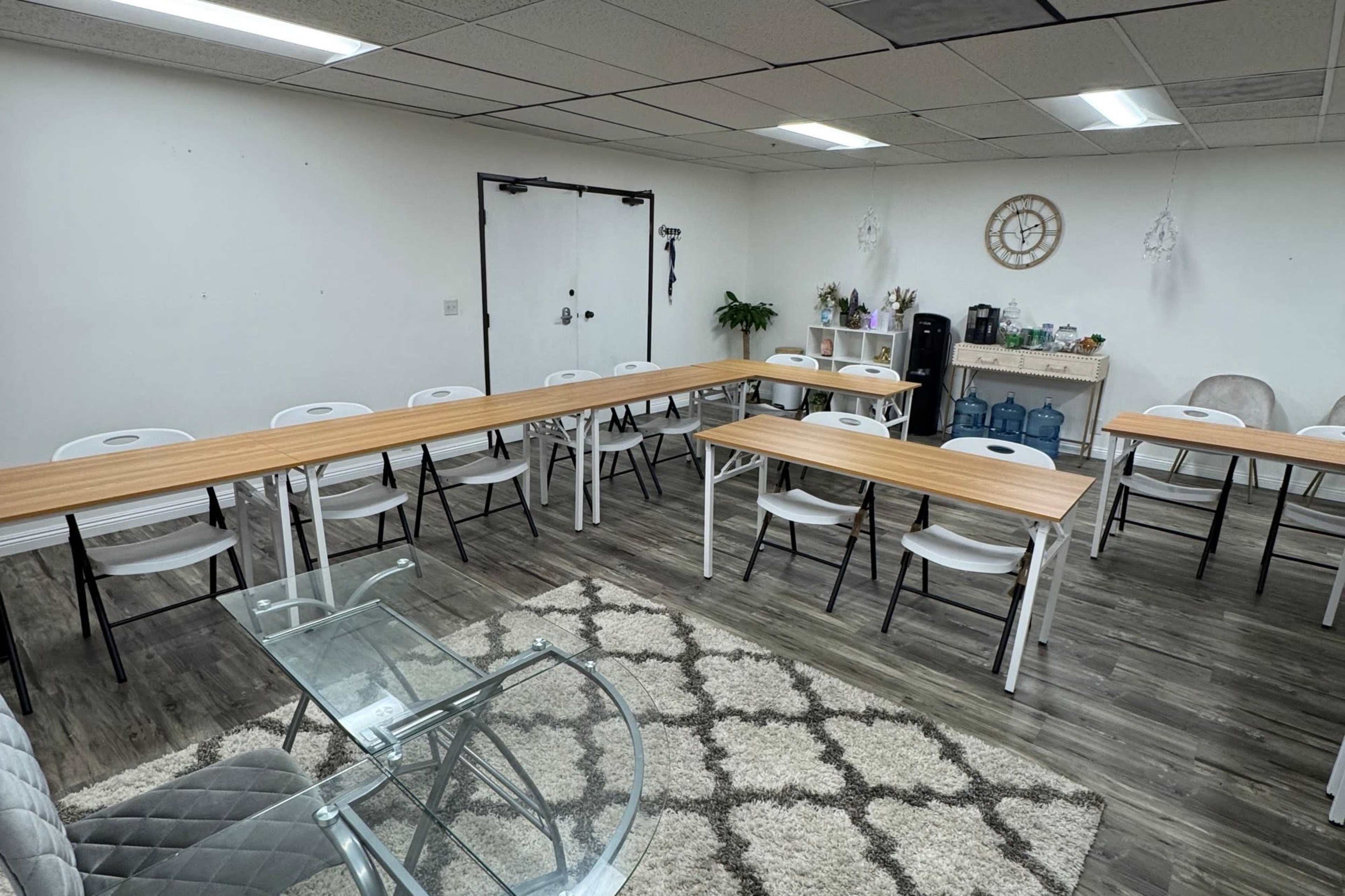 The image shows a modern meeting room with several tables arranged in a U-shape, surrounded by chairs, and featuring a decorative rug, plants, and a kitchenette in the background.