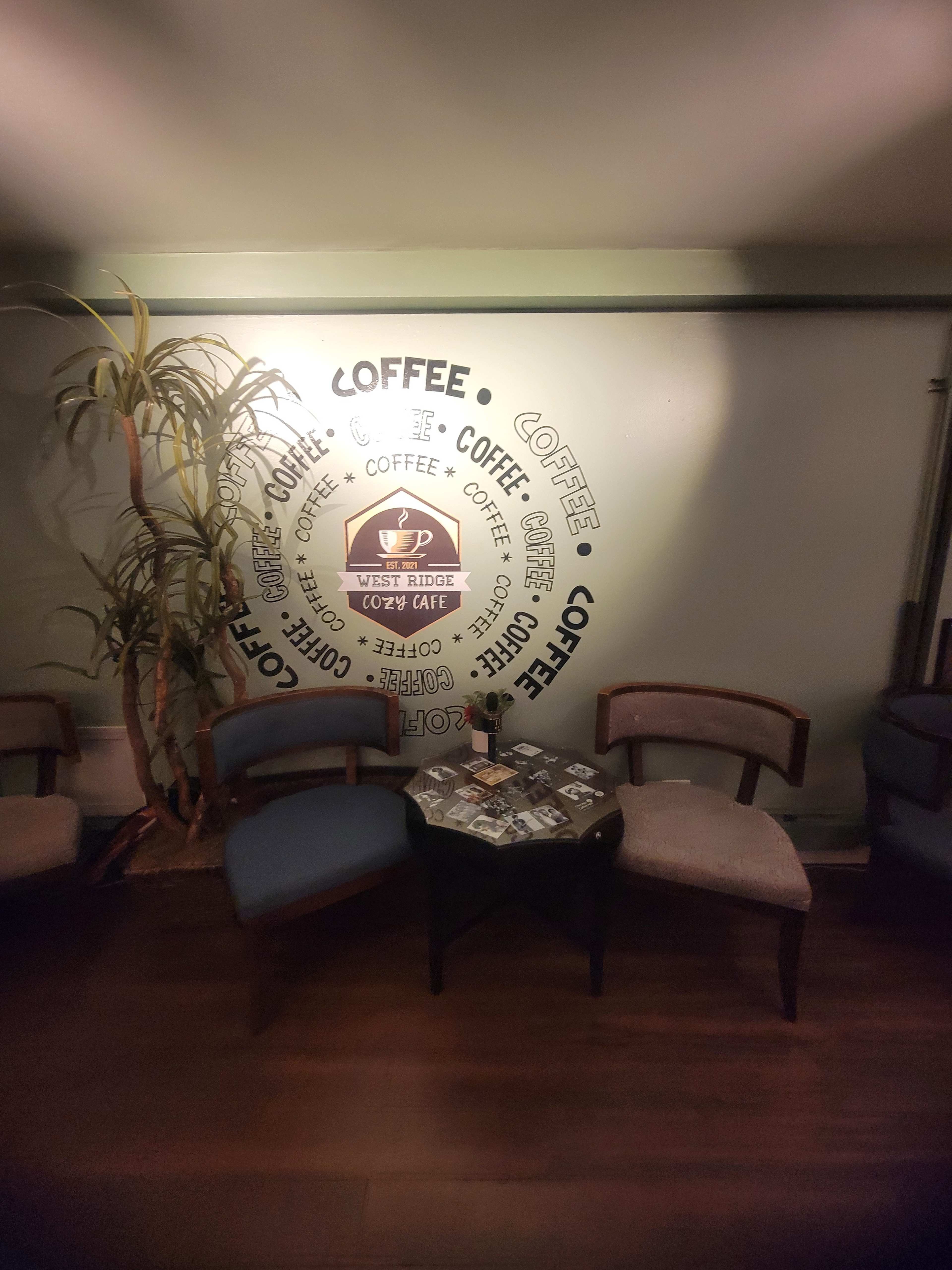 The image shows a cozy corner of a café with two chairs and a small round table against a wall decorated with a coffee-themed design.