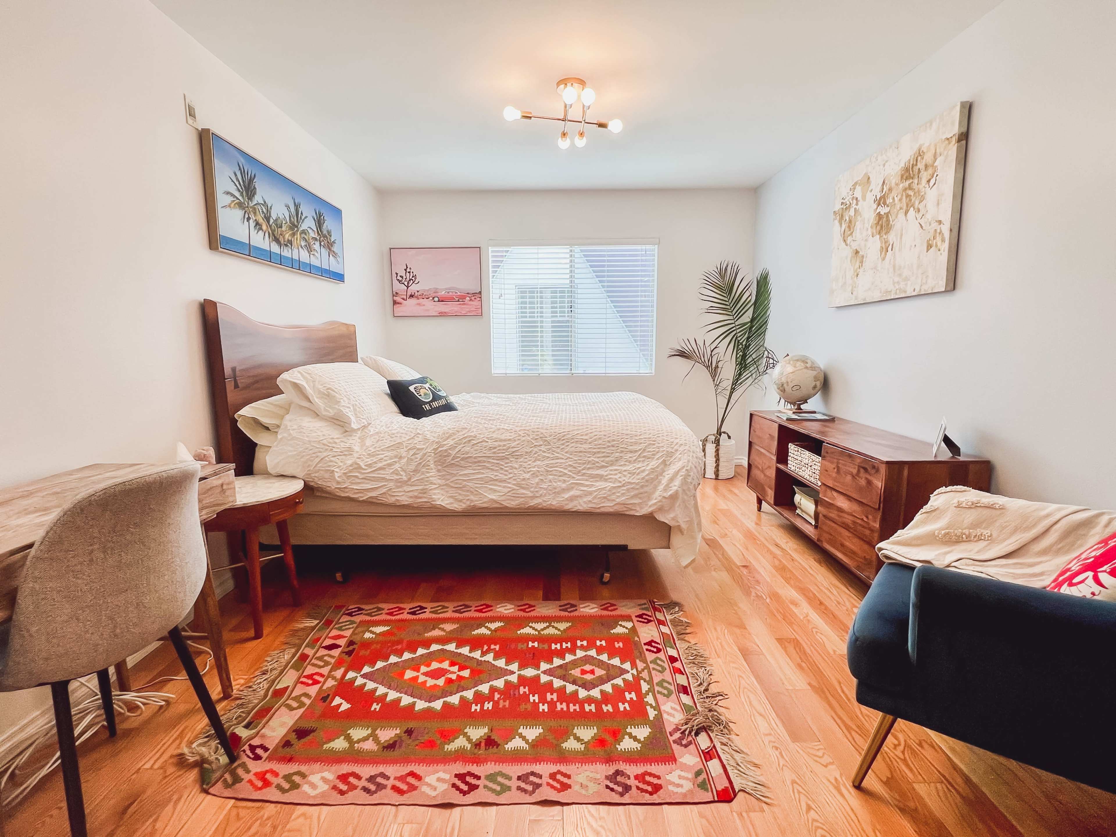 The image shows a simple bedroom with a bed, a small desk, a dark blue chair, and wooden furniture, along with a colorful patterned rug.
