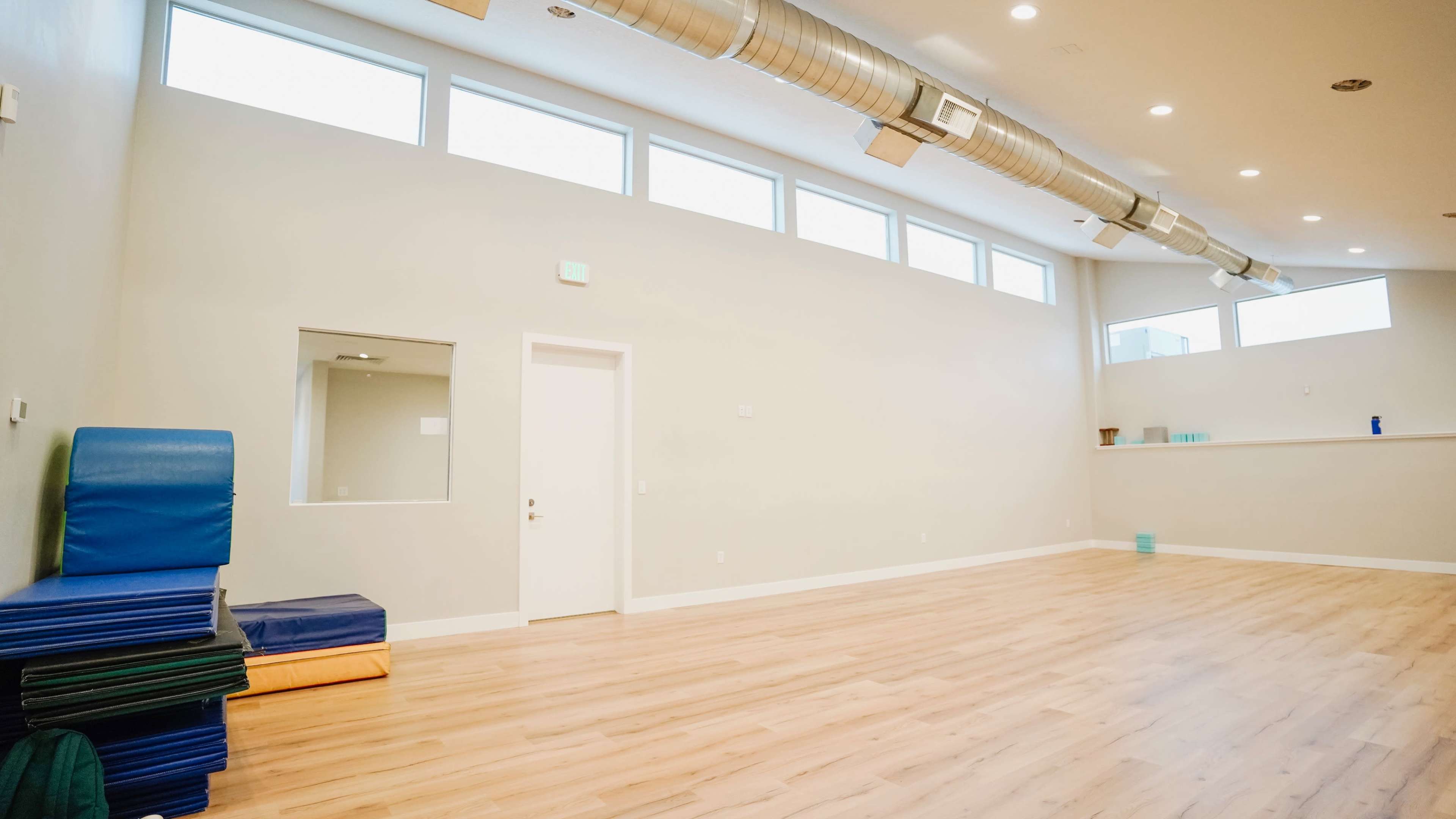 A spacious, minimally furnished room with large windows, light-colored walls, and several stacked exercise mats along one side.