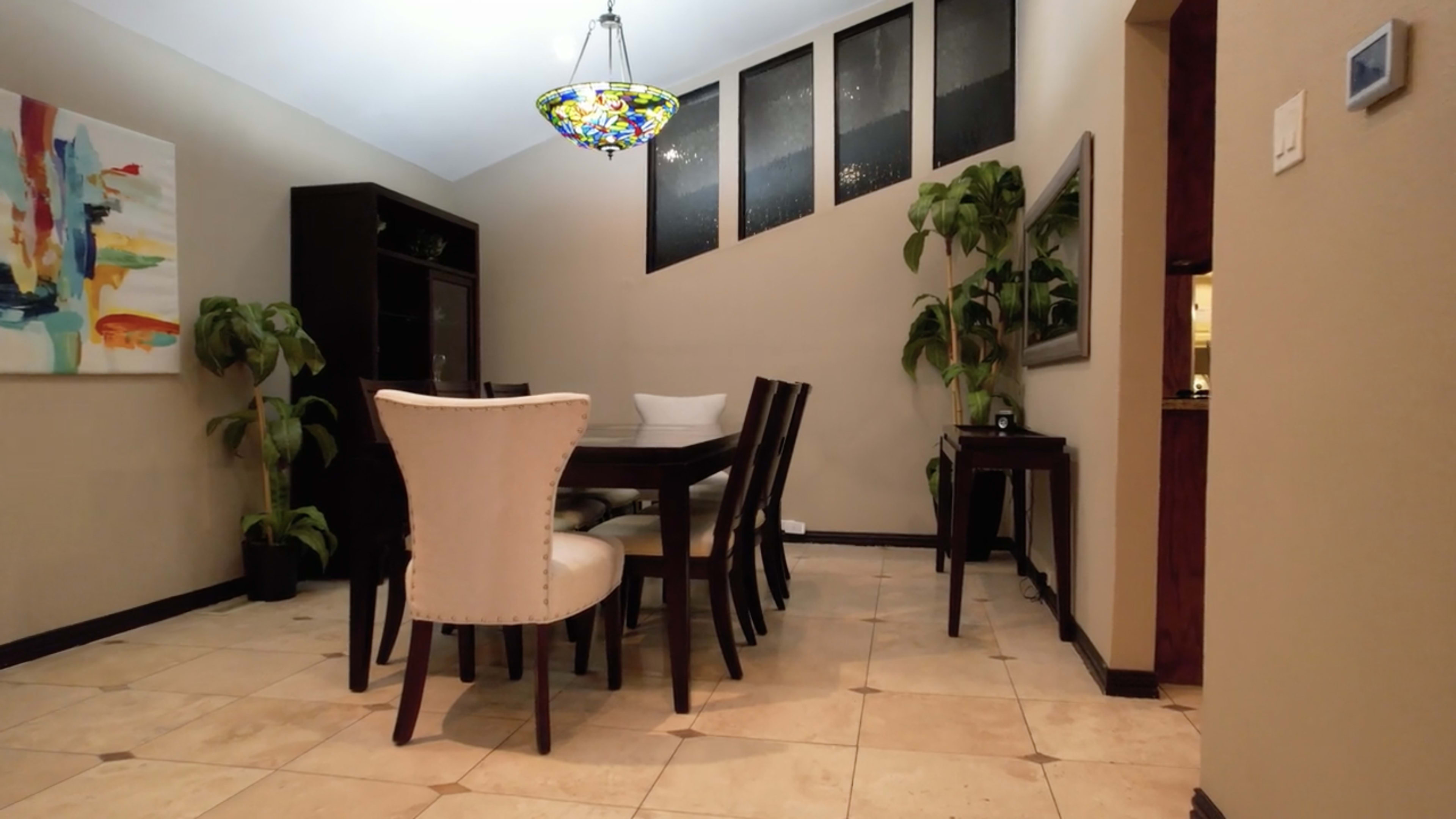 A dining room features a wooden table surrounded by chairs, with a stained glass chandelier hanging above, and plants in the corners.