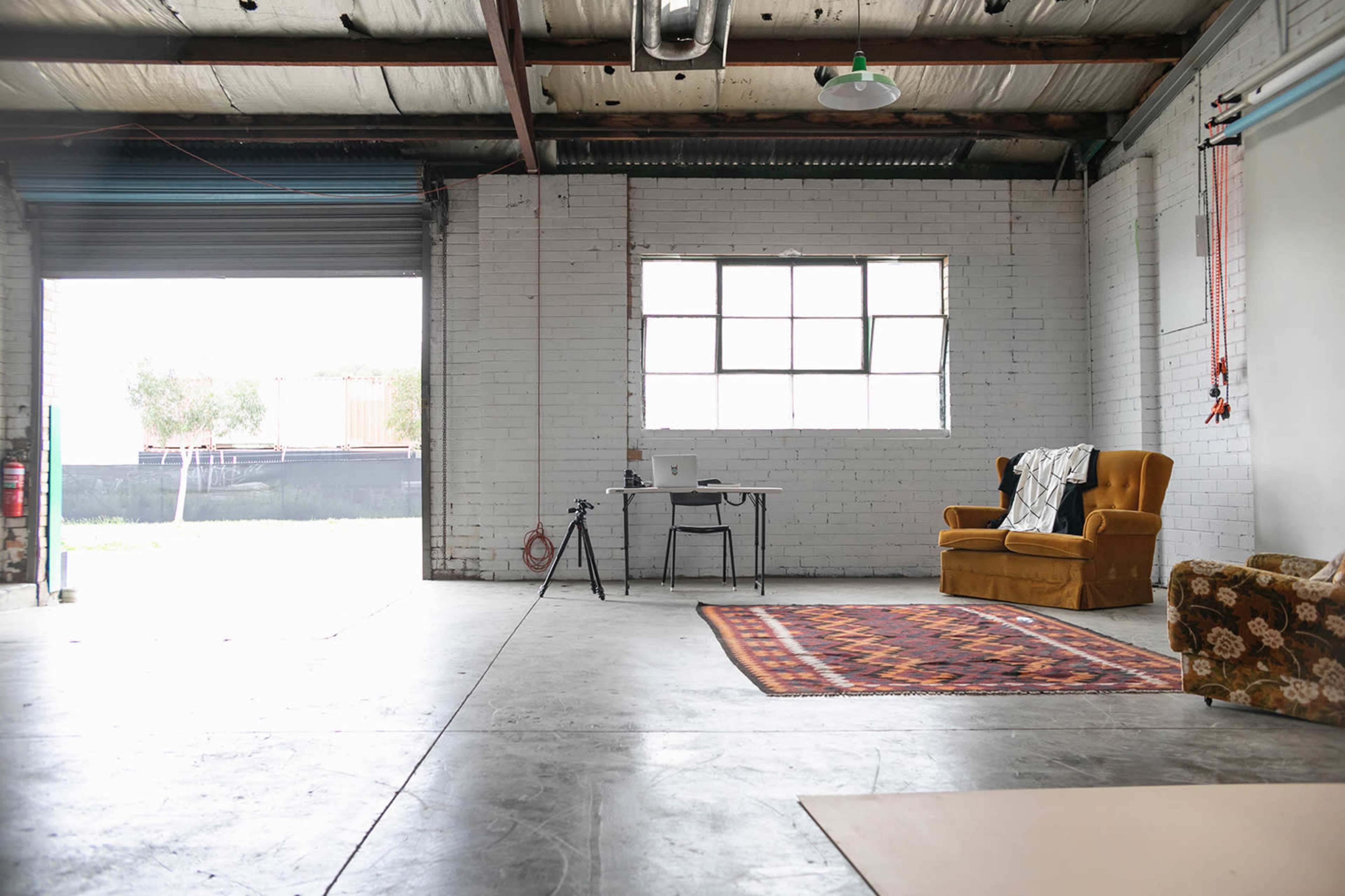 A spacious industrial room with a large window, a brown couch, a desk with a laptop, and a tripod on the floor.