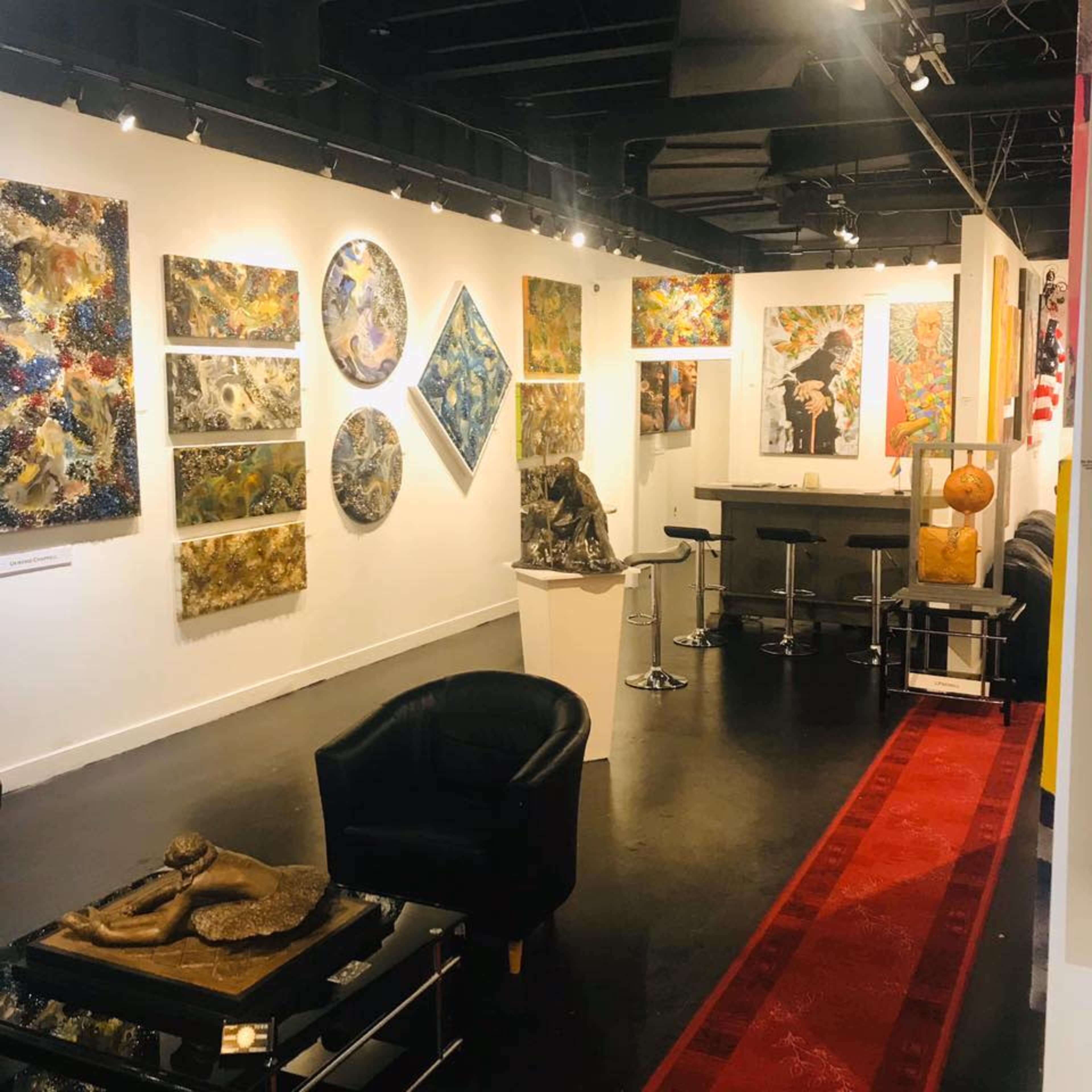 A modern art gallery featuring various colorful paintings and sculptures displayed on white walls, with a seating area consisting of black chairs and a bar-style table.