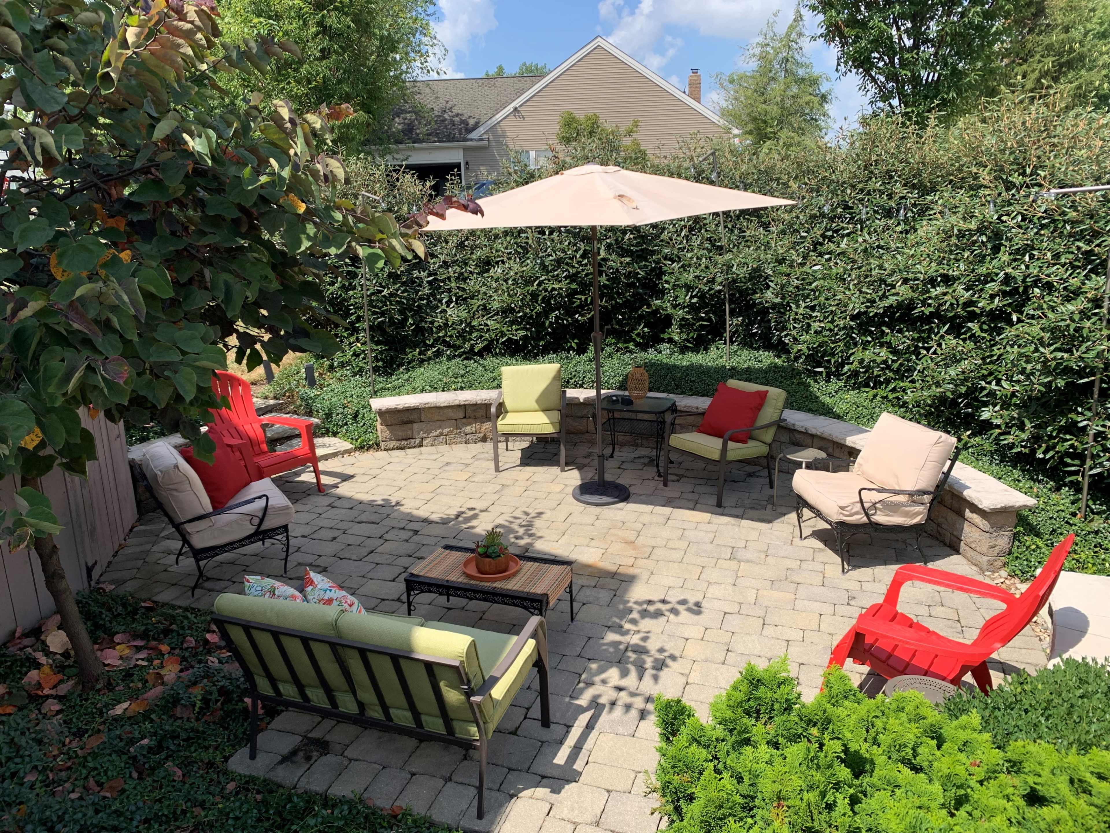 A circular stone patio features various seating options, an umbrella, and a small table surrounded by lush greenery.