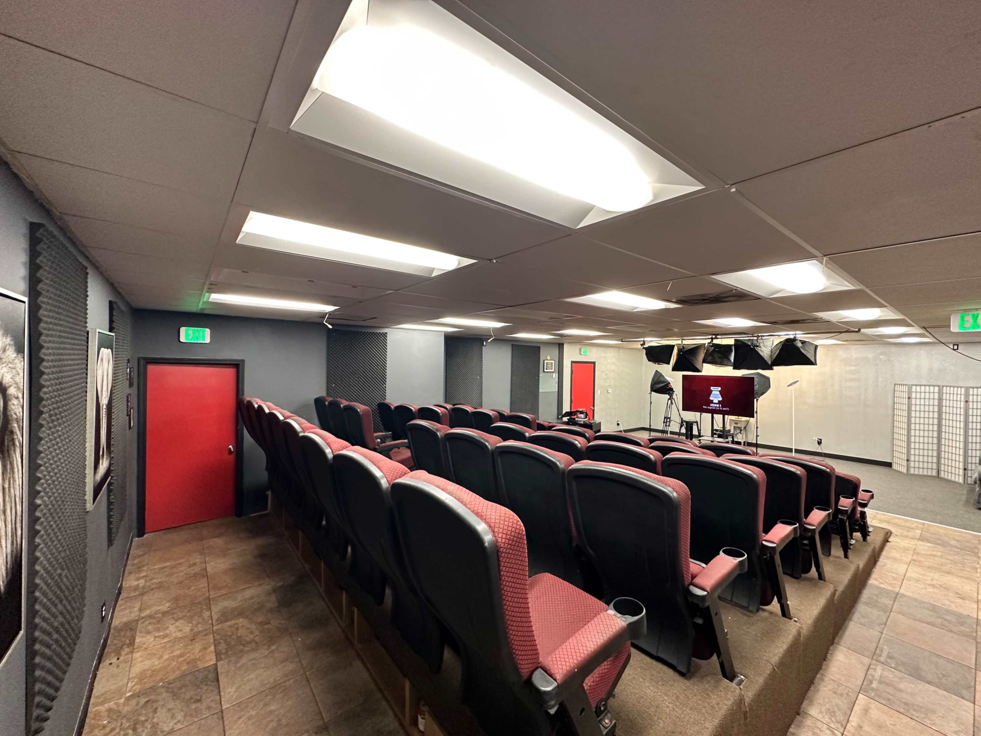An indoor auditorium with tiered seating, featuring red chairs and a small stage at the front.