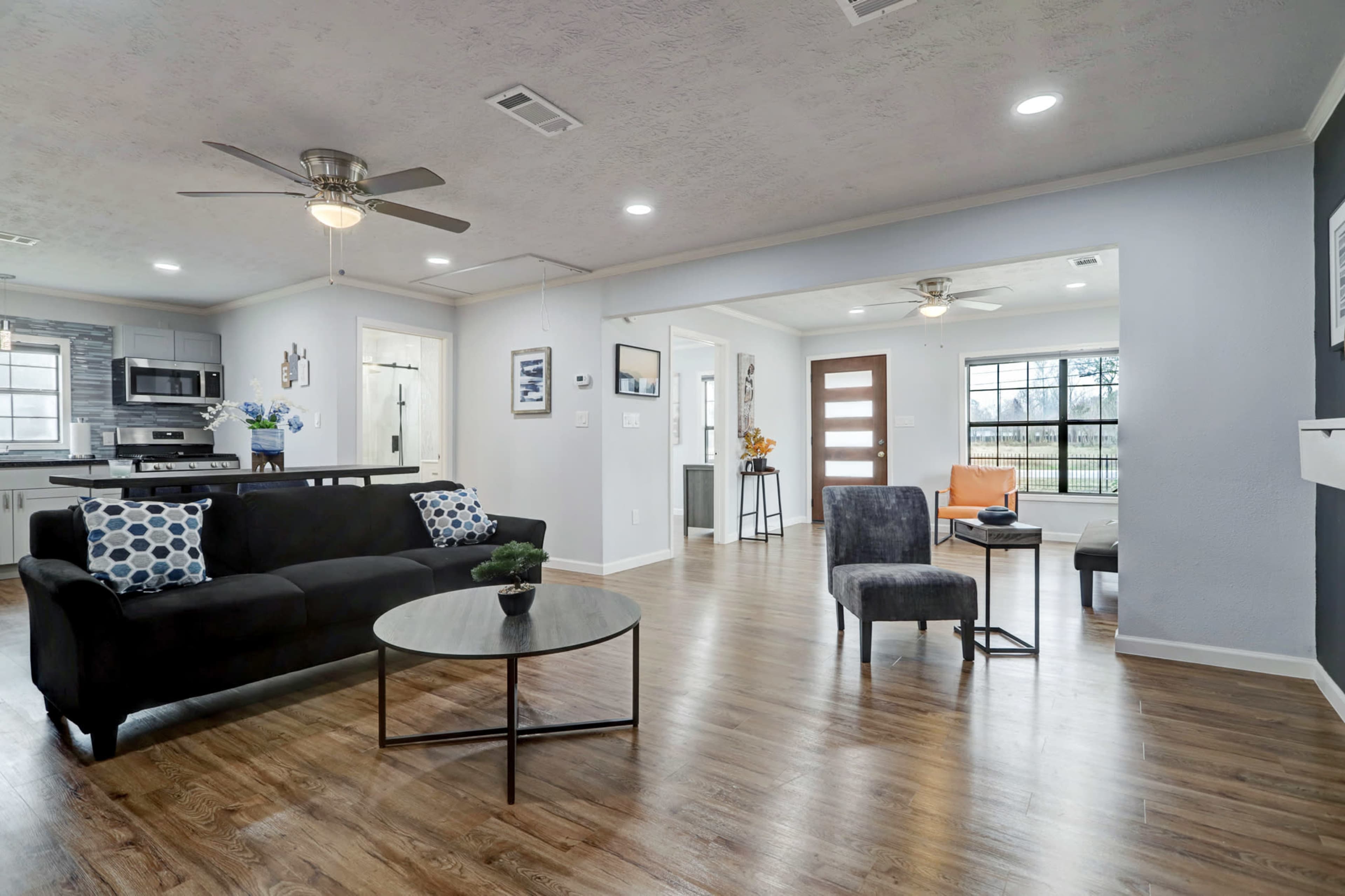 A modern living room with a black couch, a round coffee table, and a view into a bright kitchen area, featuring wood flooring and large windows.