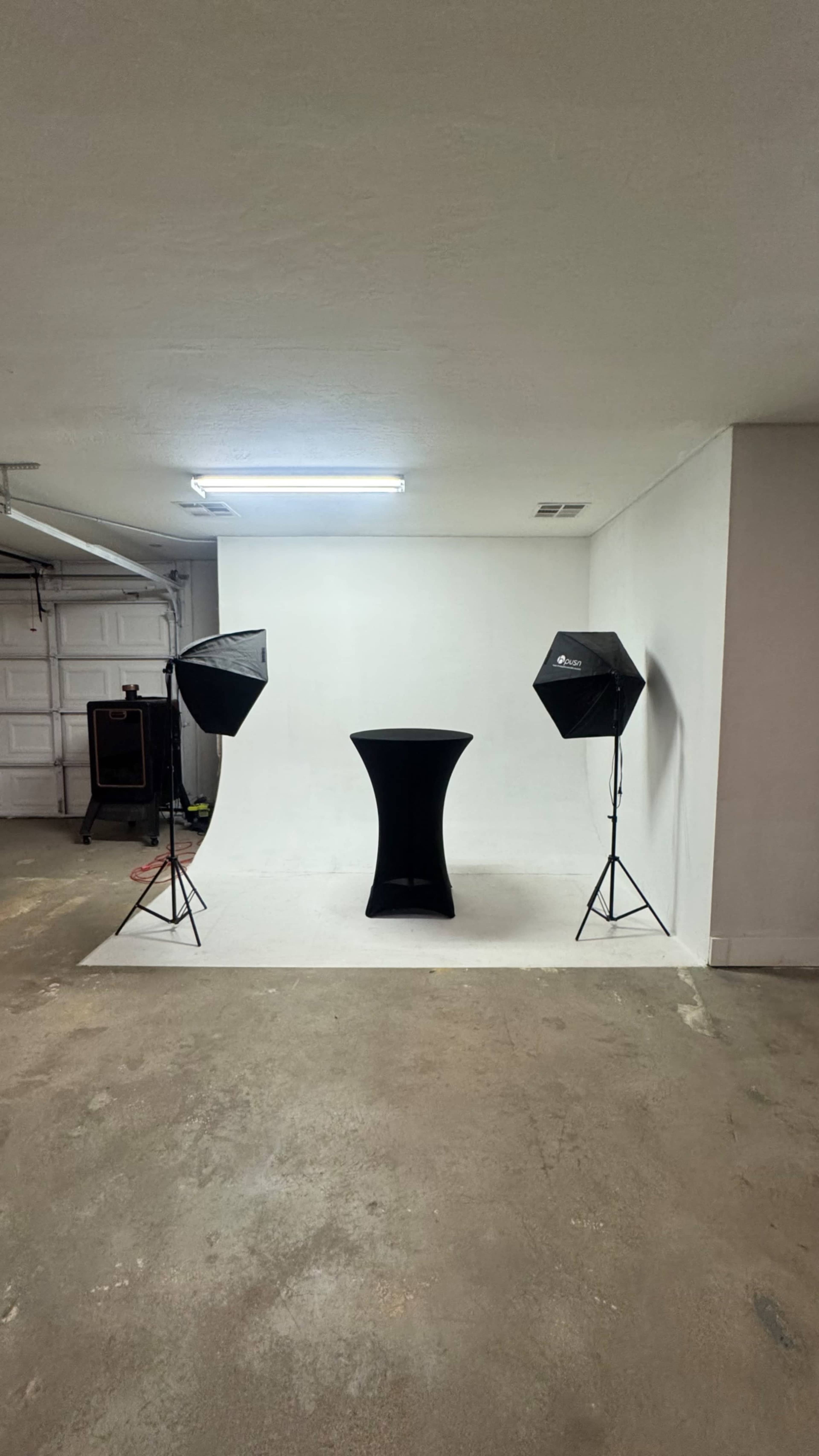 The image shows a plain studio setup featuring a black table in front of a white backdrop, with two softbox lighting fixtures positioned on either side.