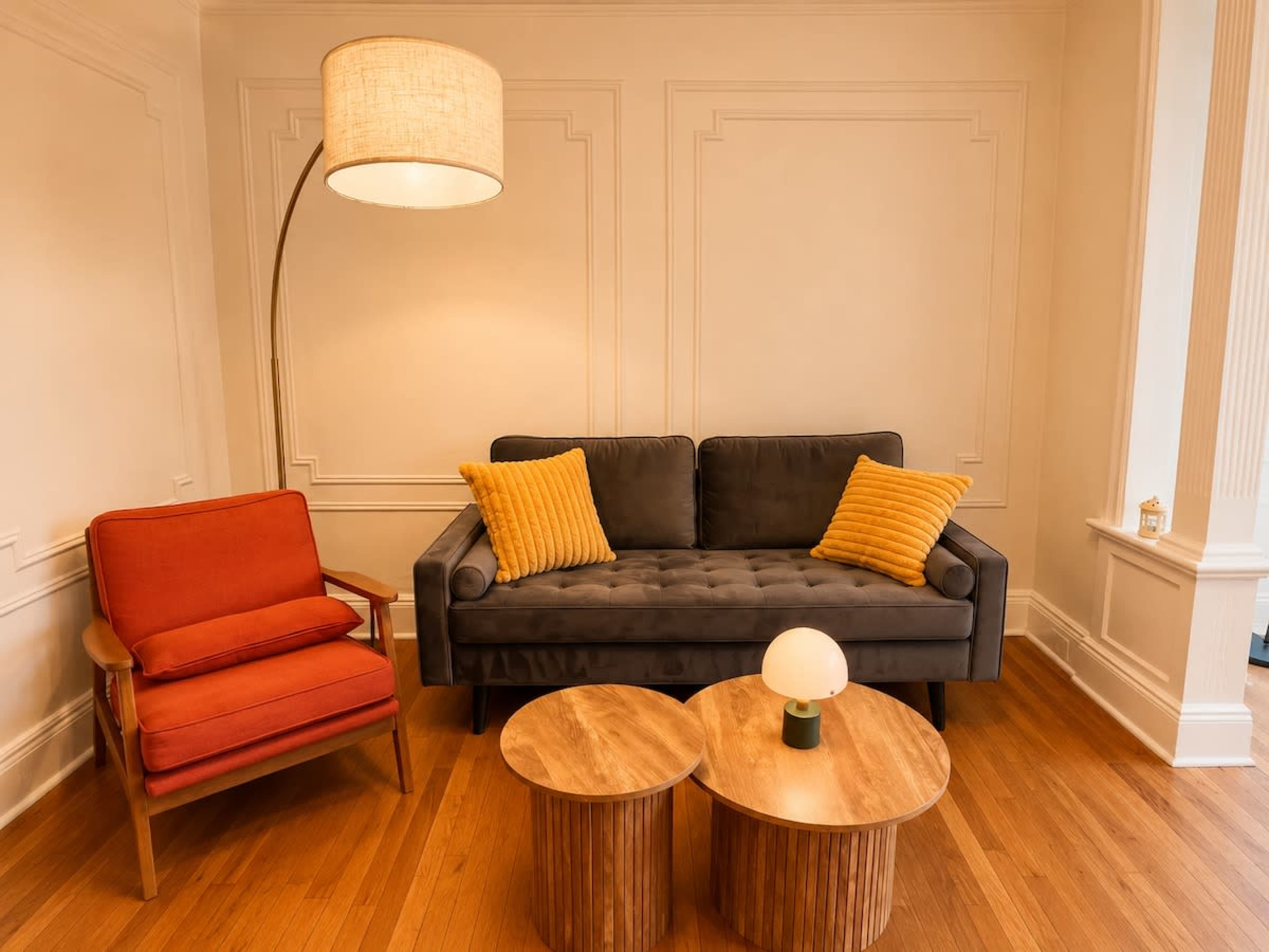 The image shows a modern living room setup with a dark gray sofa, an orange armchair, two wooden coffee tables, and a standing lamp.