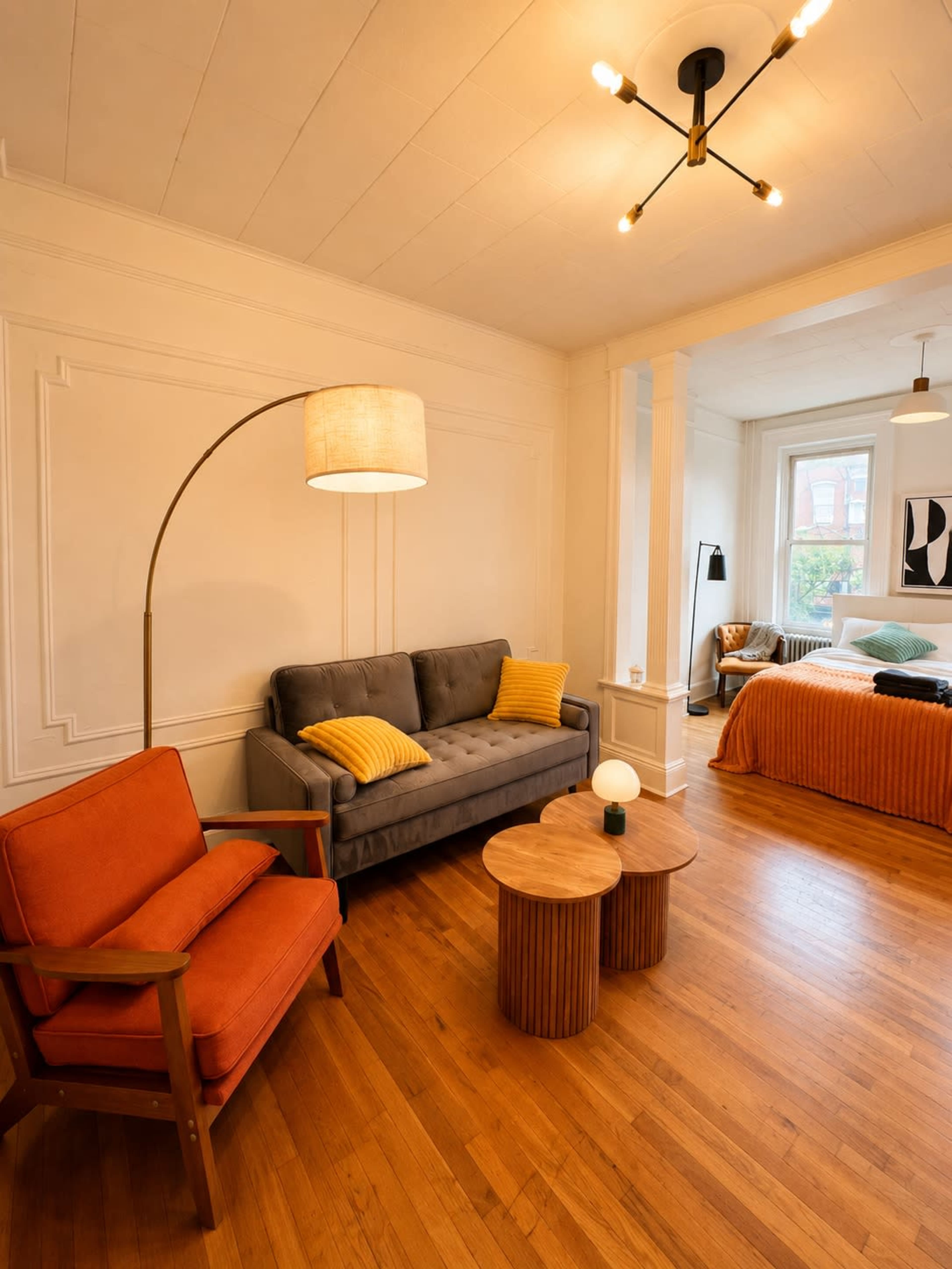 The image shows a cozy living space featuring a gray sofa and an orange armchair, with two round wooden coffee tables and a bed in the background.