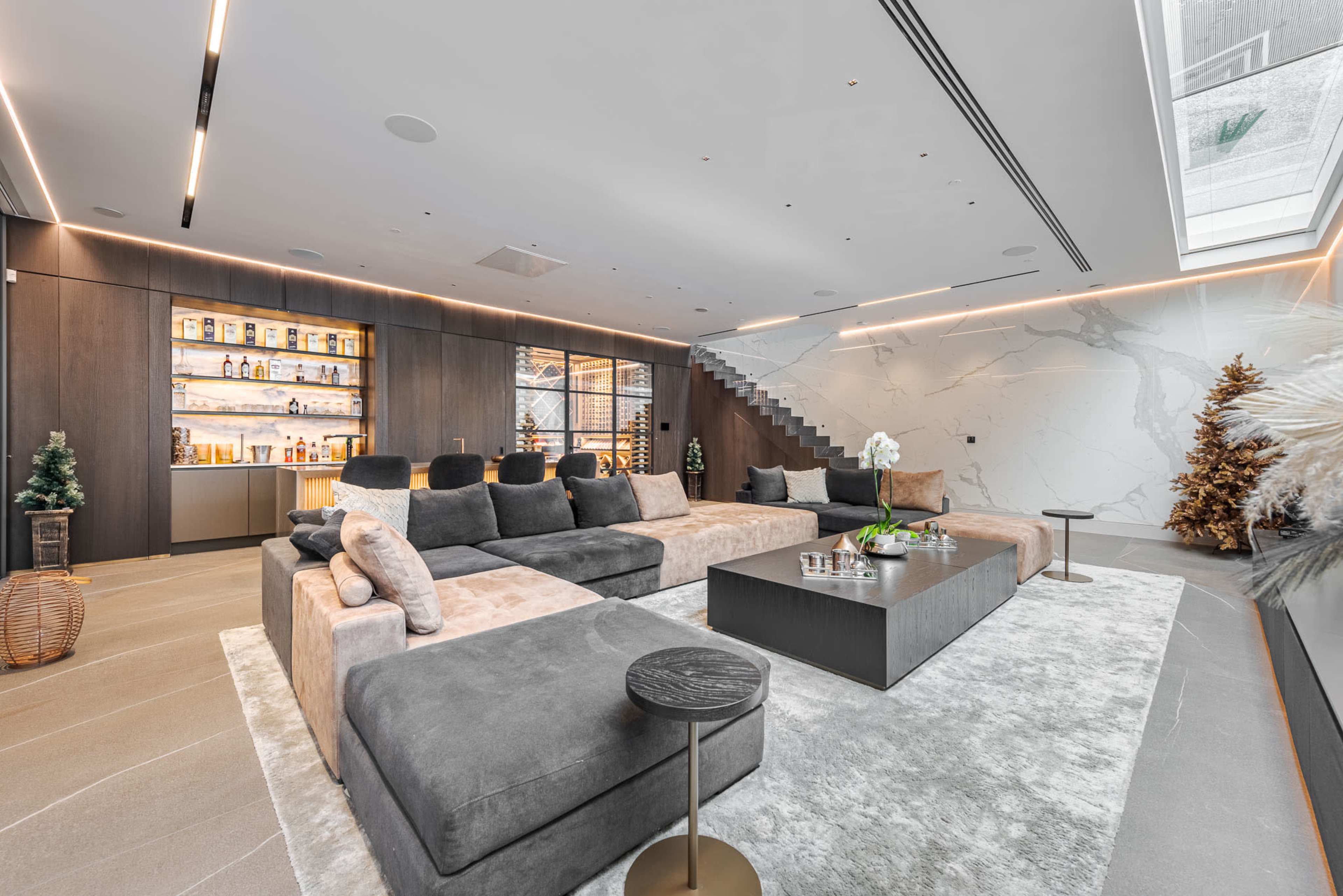 The image shows a modern living room featuring a large sectional sofa, a coffee table, and a wall with shelves displaying various bottles, all set against a backdrop of stylish decor and large windows.