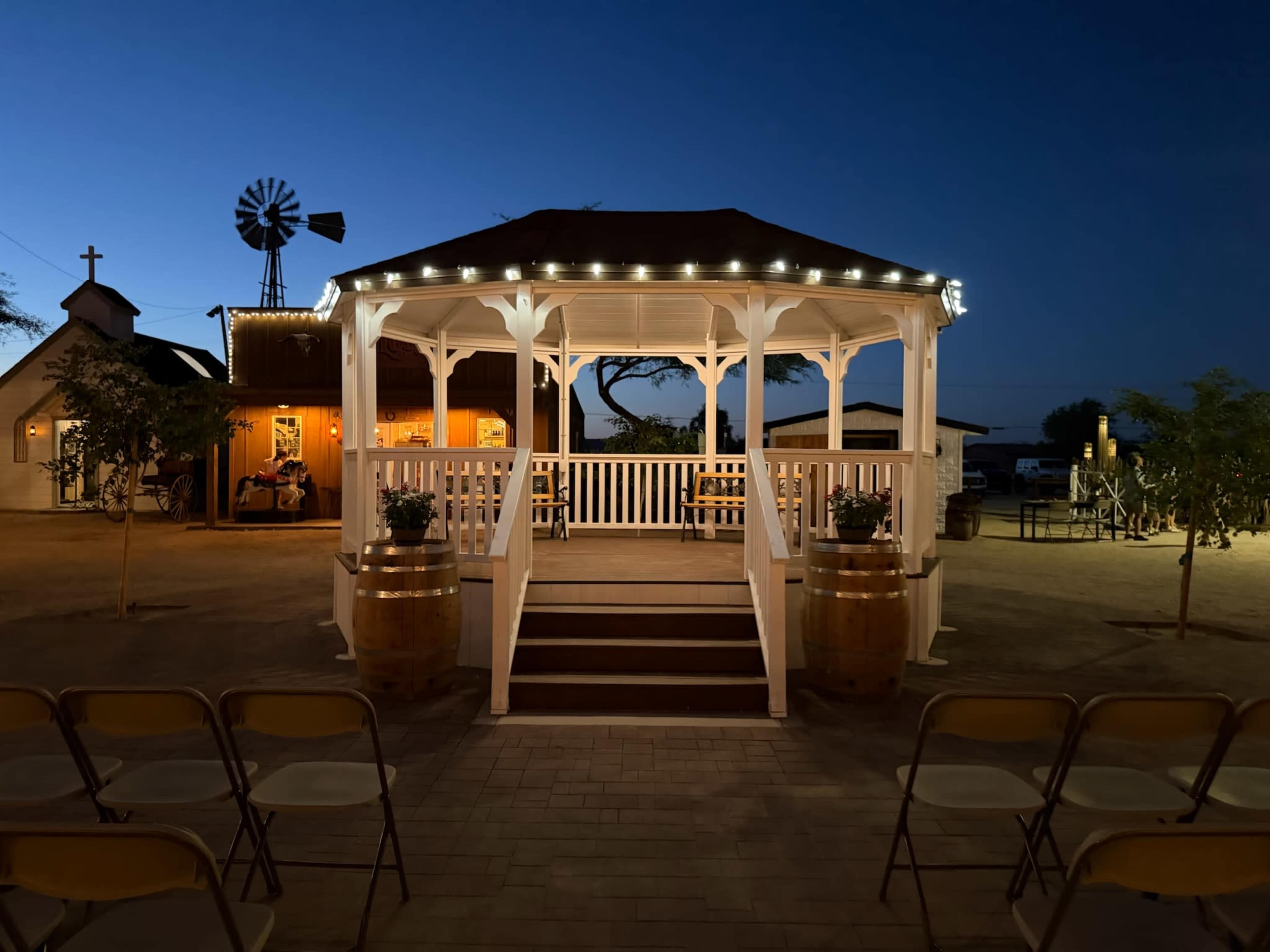 A gazebo is lit with string lights and surrounded by empty chairs in a rustic outdoor setting at dusk.