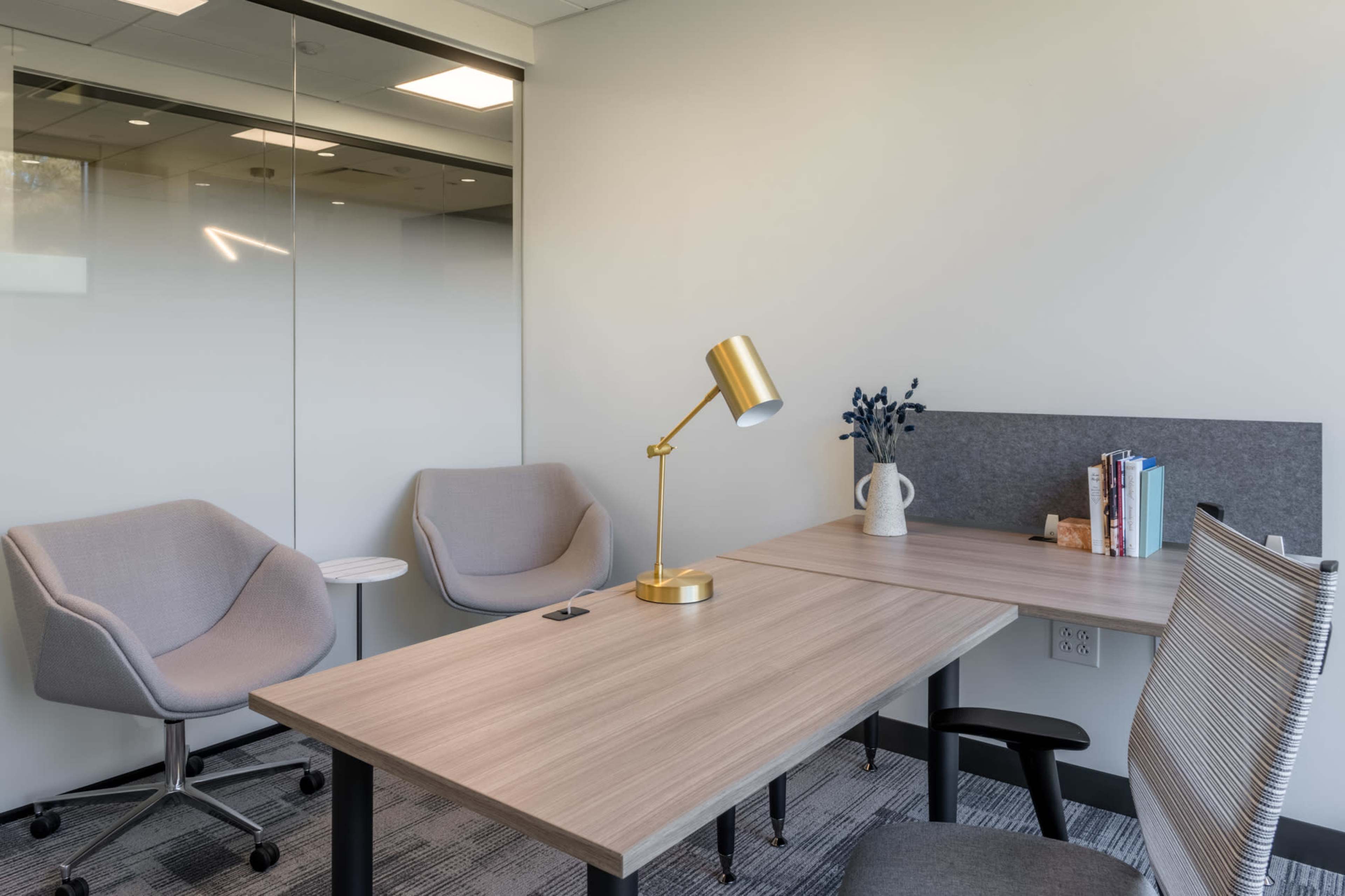 A modern office space features a wooden desk with a gold lamp, two gray chairs, and a small table, along with a book collection and decorative items arranged neatly on the desk.