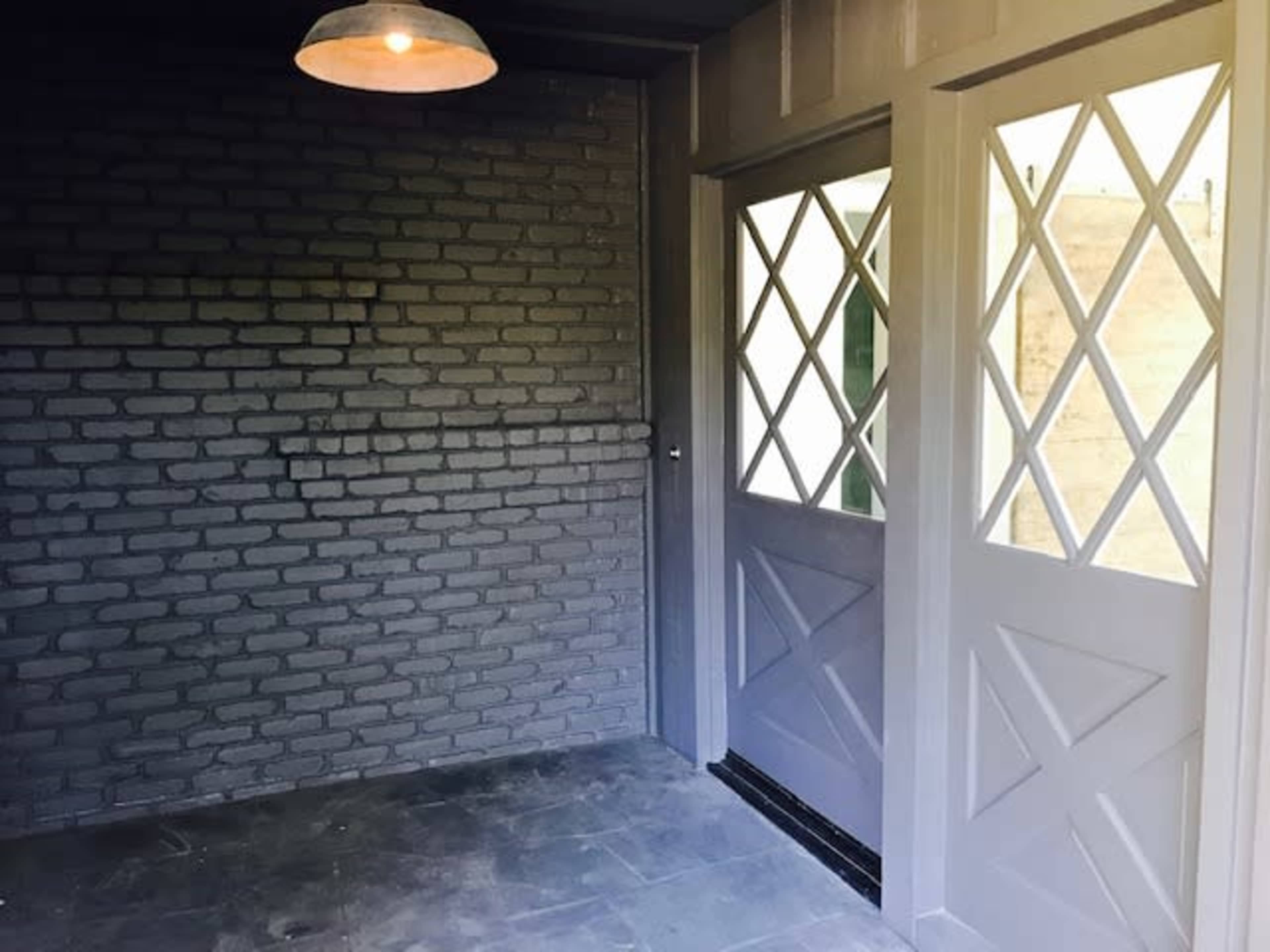 The image shows a room with gray brick walls, a concrete floor, and double doors with diamond-patterned glass panels allowing natural light to enter.