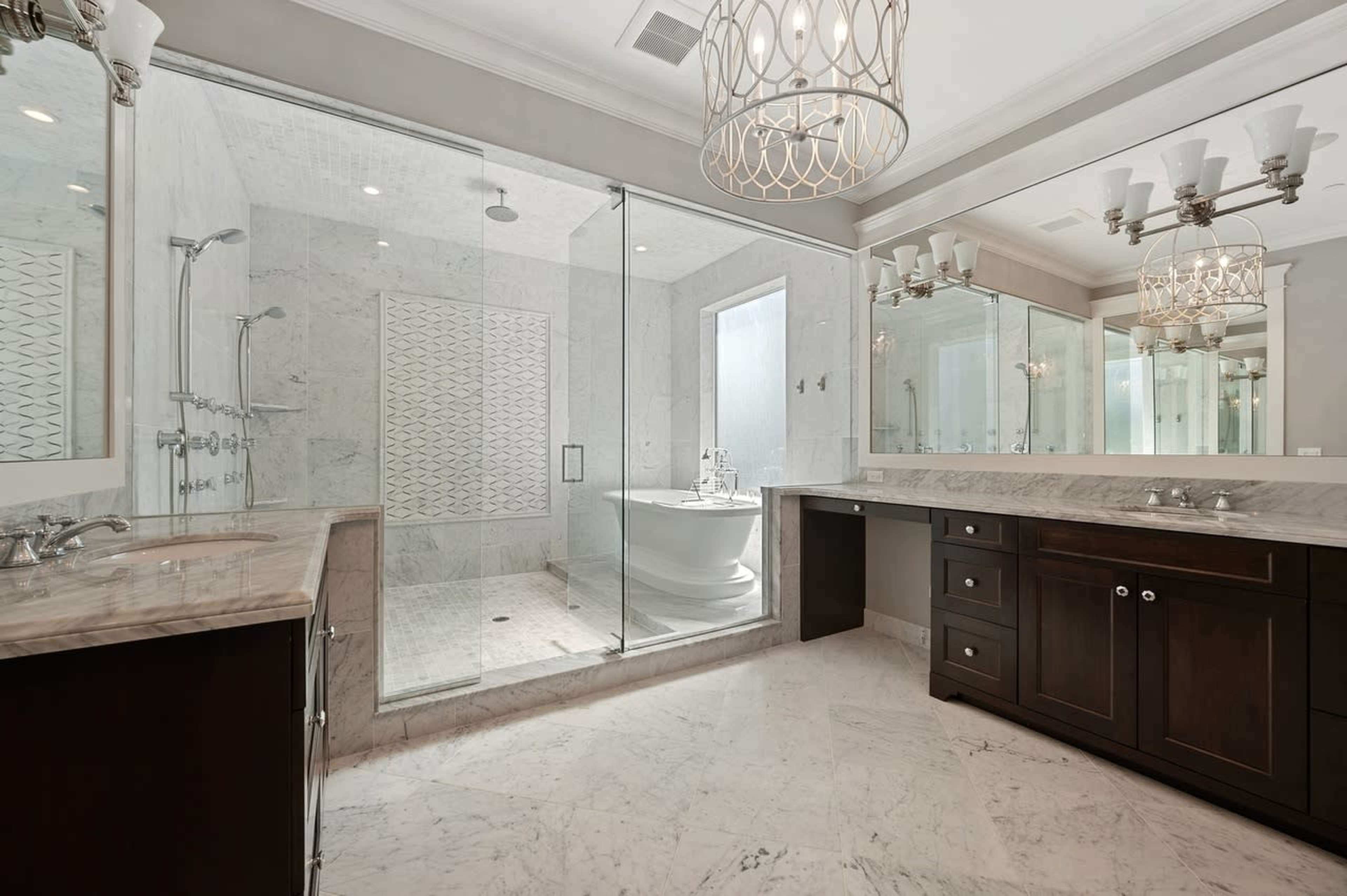 A modern bathroom featuring a glass-enclosed shower, a freestanding bathtub, dual vanities with dark wood cabinetry, and marble flooring.