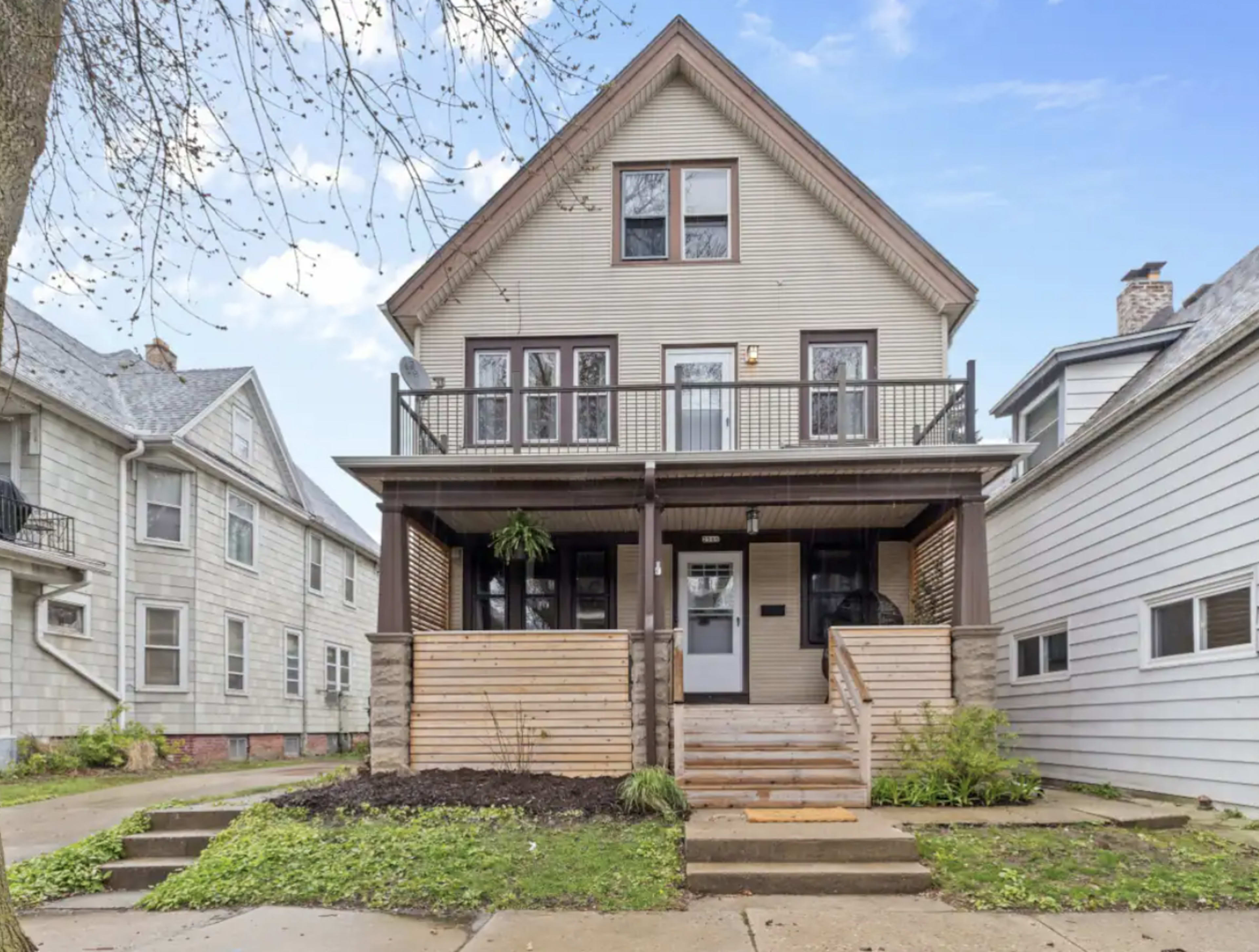 A three-story house with a front porch, wooden railings, and landscaped steps is situated on a tree-lined street.
