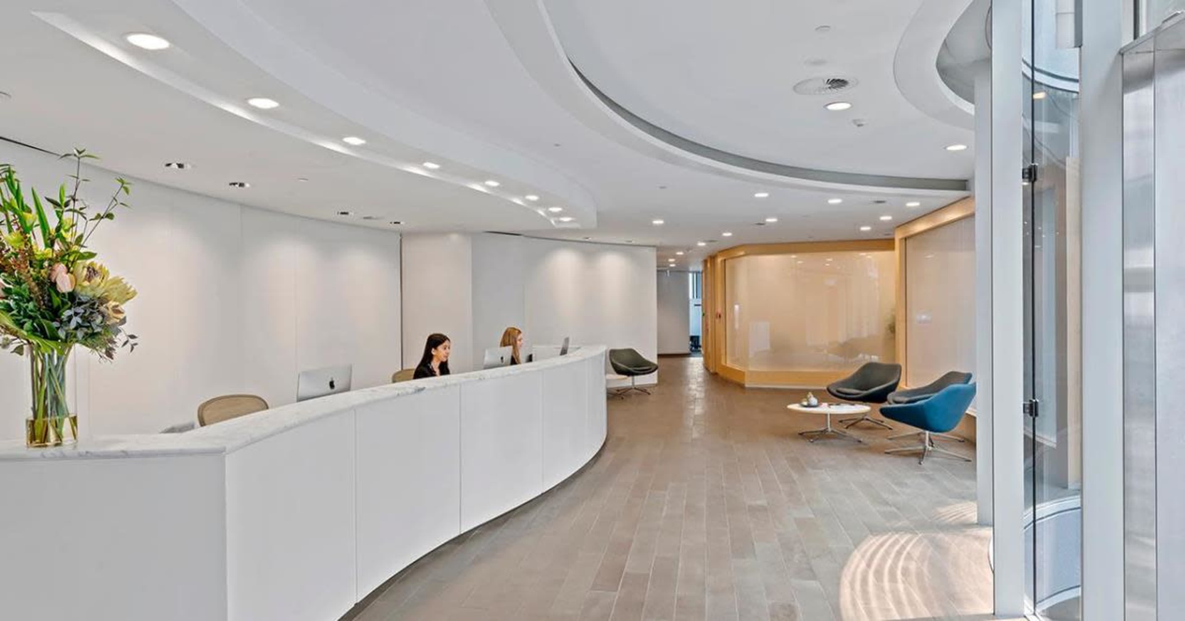 A modern reception area featuring a curved white front desk, two women seated at computers, and contemporary seating arrangements in a well-lit space.