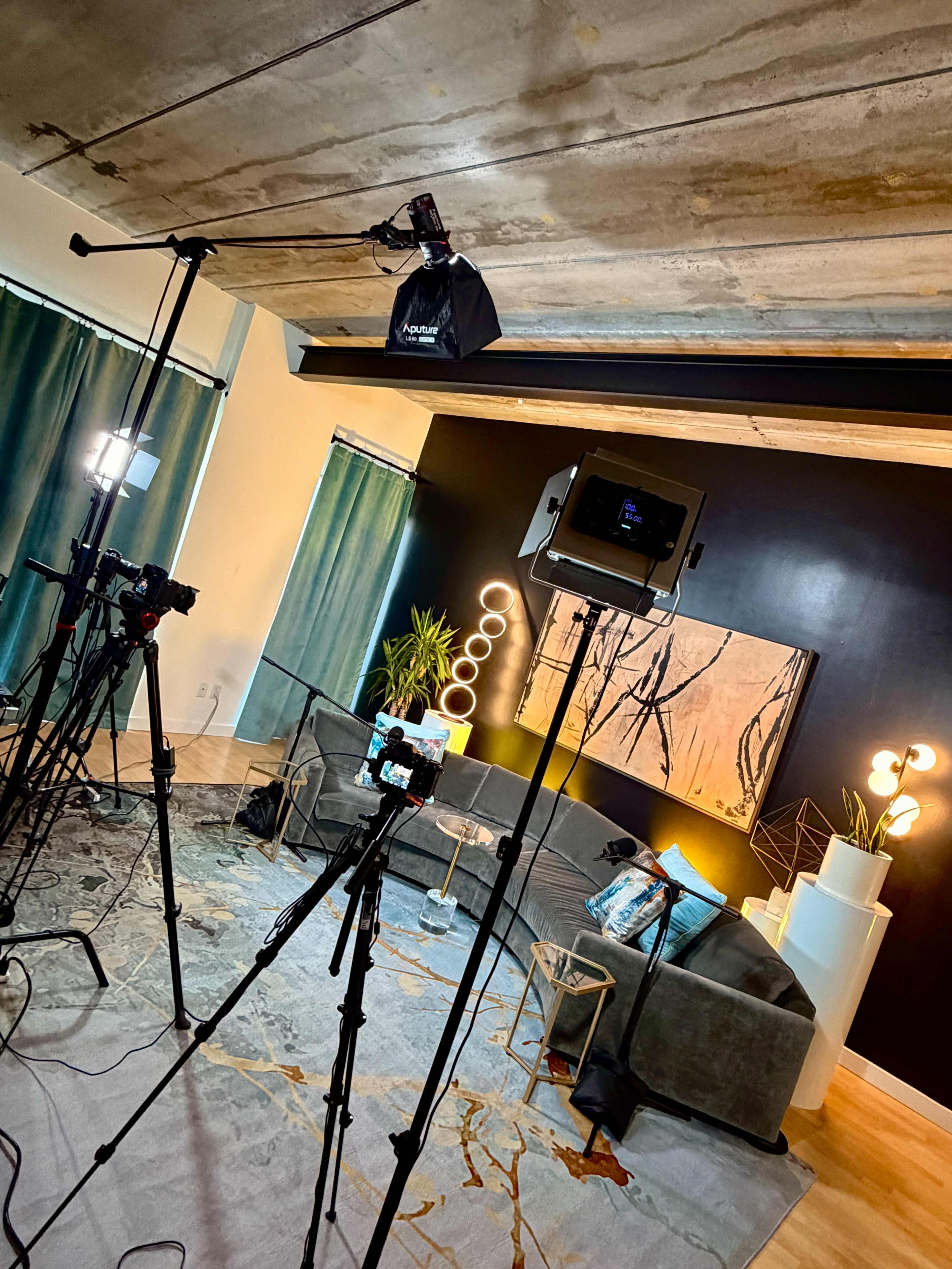 The image shows a well-lit studio setup with multiple cameras and lighting equipment arranged around a modern living room featuring a grey couch, decorative plants, and abstract wall art.