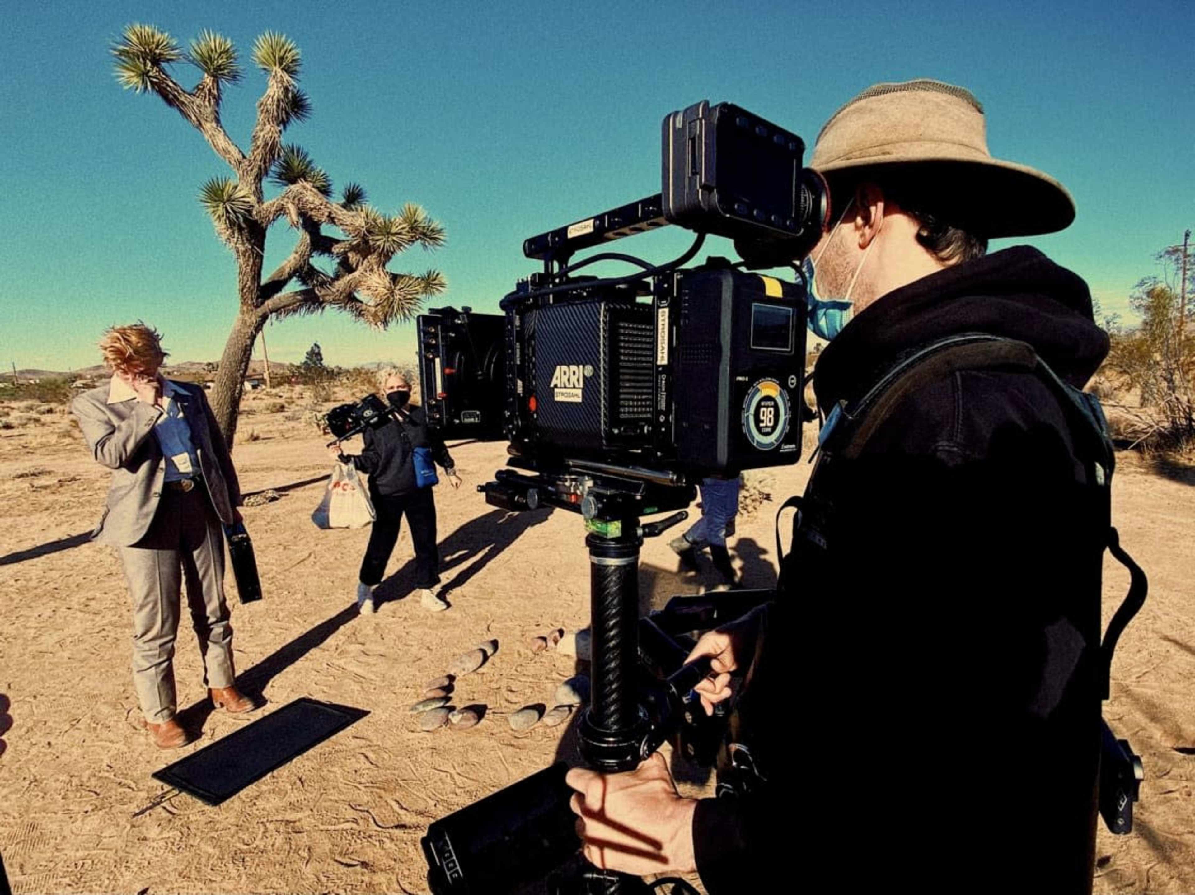 A film crew is set up in a desert landscape, with a woman in formal attire standing in front of the camera while crew members adjust equipment.